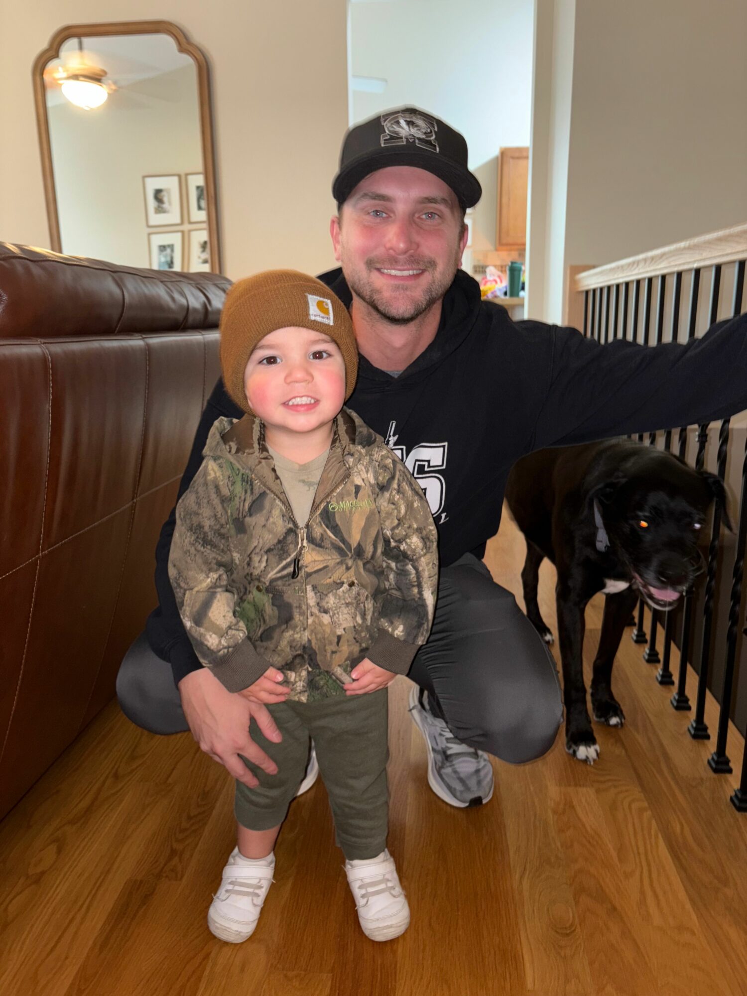 A man and a young child pose with a black dog indoors, smiling, with a mirror and framed pictures in the background.