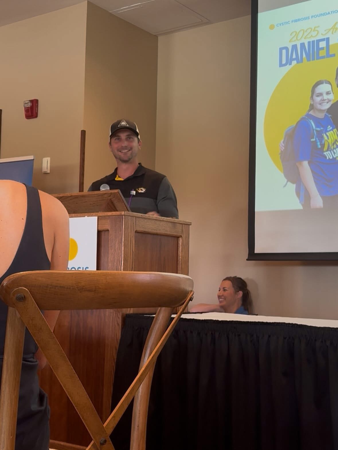 Man standing at podium, smiling, wearing a cap and dark clothing, in a room with a presentation screen showing a young person with a backpack.