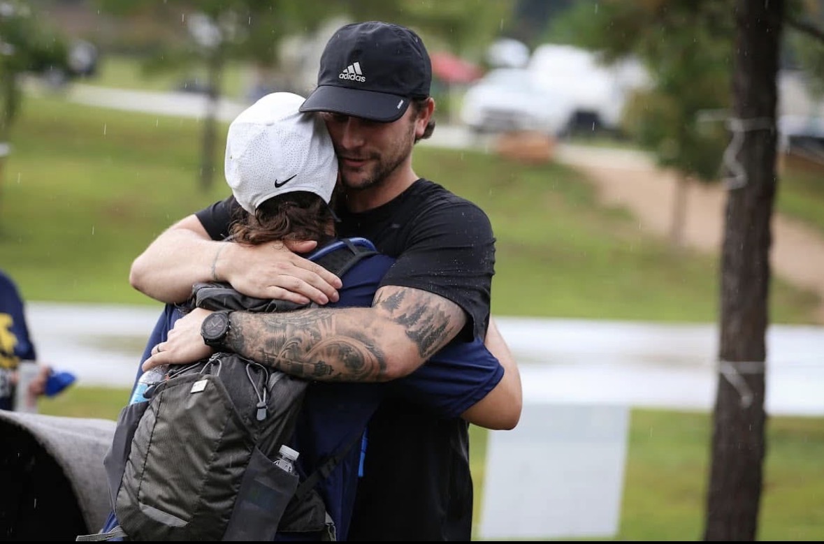 Man hugging a child outdoors with trees and a road in the background.