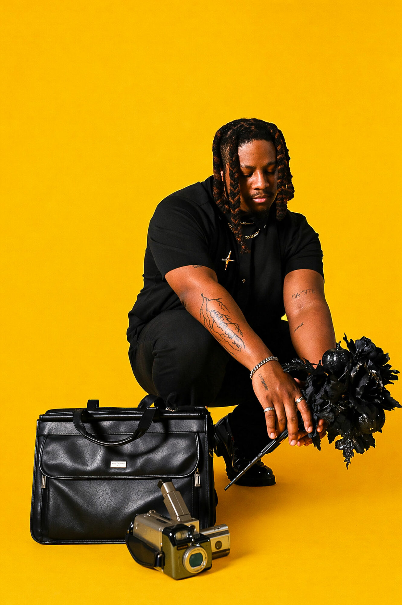 Person with dreadlocks crouching, holding a black object, with a black bag and camera on yellow background.