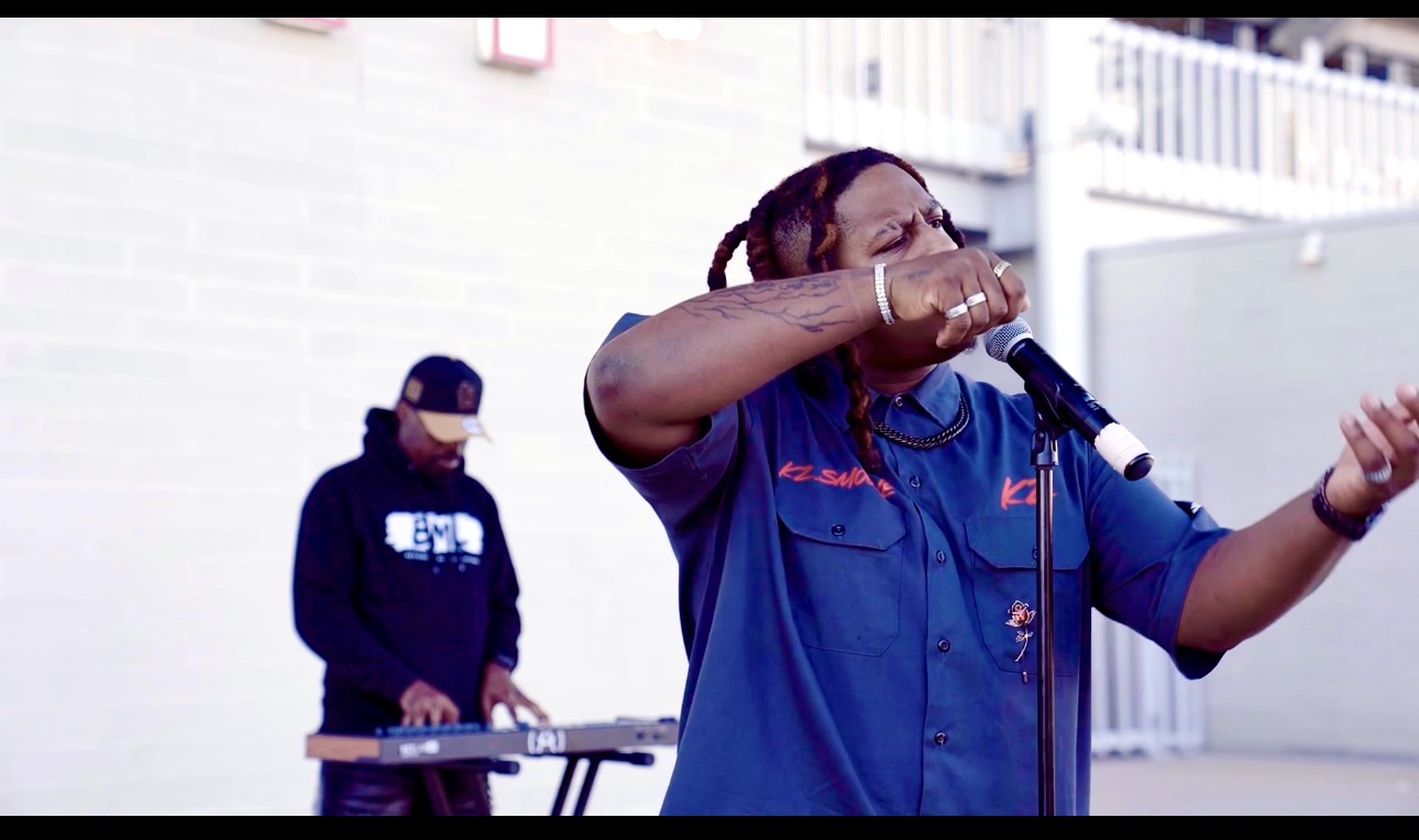 Person singing into a microphone with a DJ in the background outdoors, wearing a blue shirt, gesturing with hand