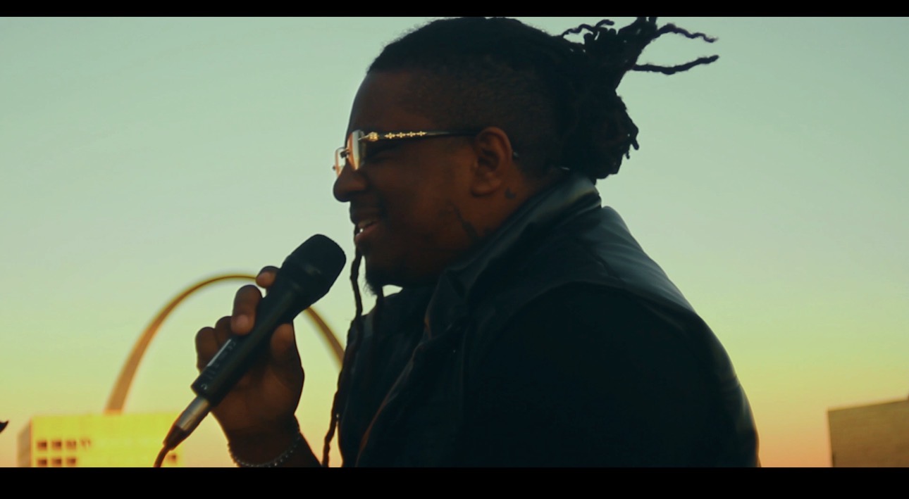 Person with dreadlocks and glasses speaking into a microphone outdoors during sunset, facing left.