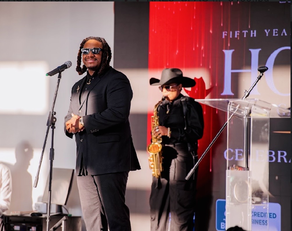 Two people on stage, one speaking into a microphone, the other playing a saxophone, with a red and black background.