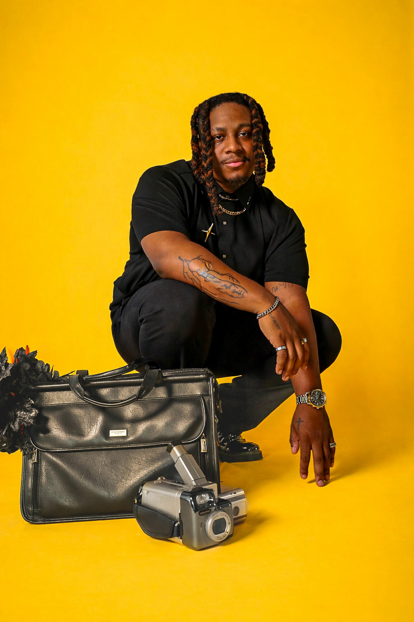 Person crouching with a black bag and camera on a yellow background, wearing black clothing and jewelry.