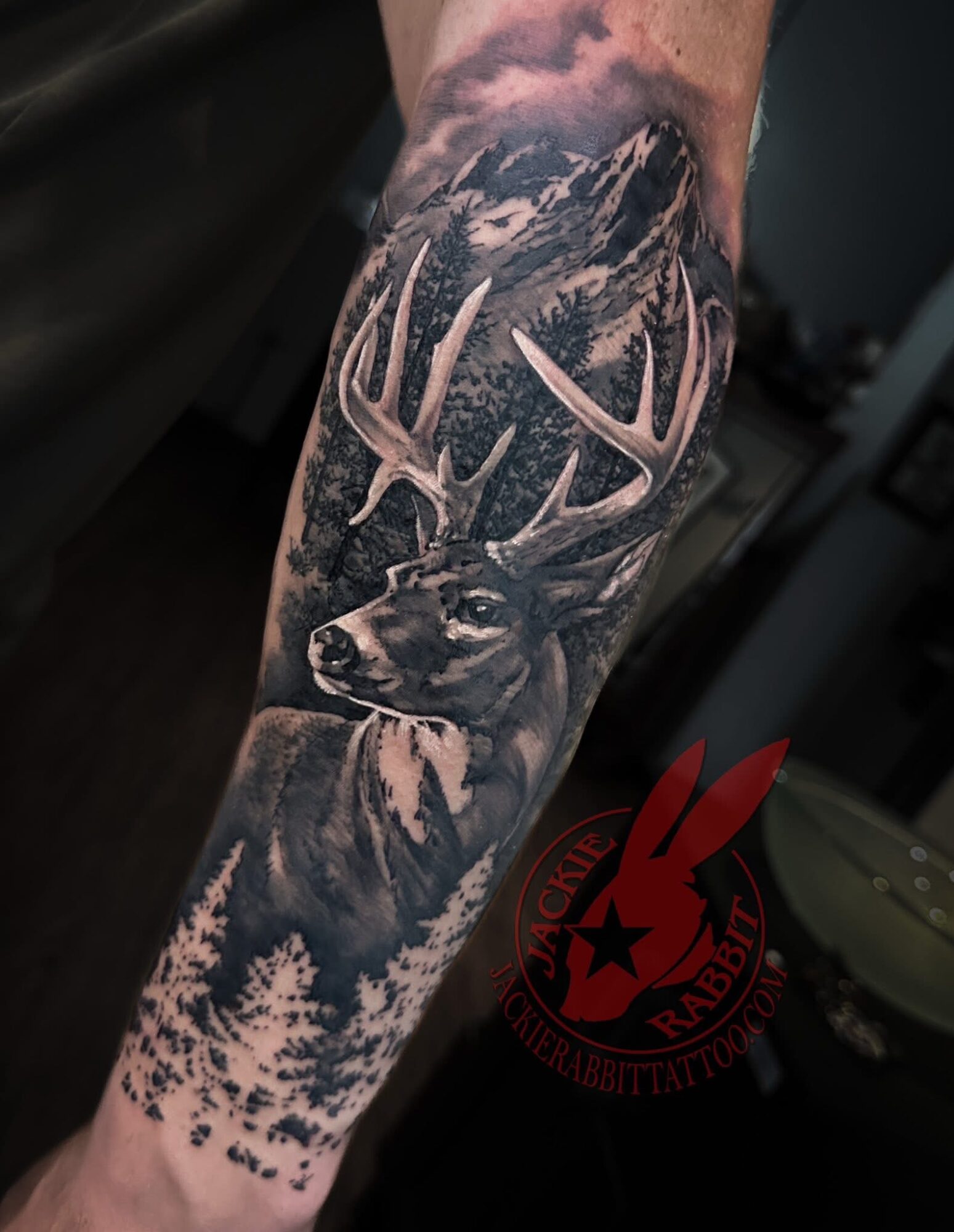 Black and gray tattoo of a deer with antlers, mountain landscape, and trees on a person's arm.