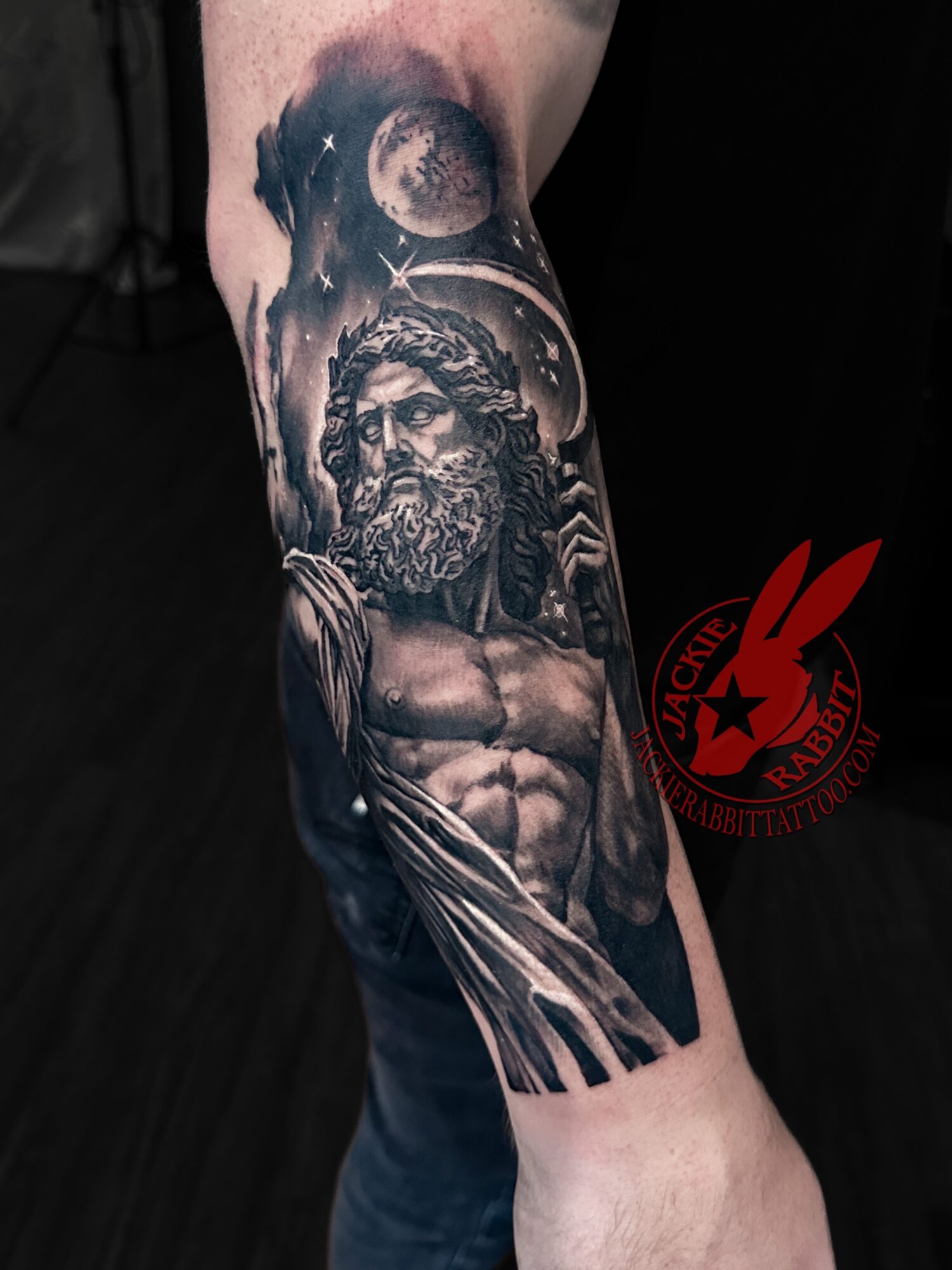Black and gray tattoo of a muscular man with a beard, wearing a crown, with celestial elements above, on arm.