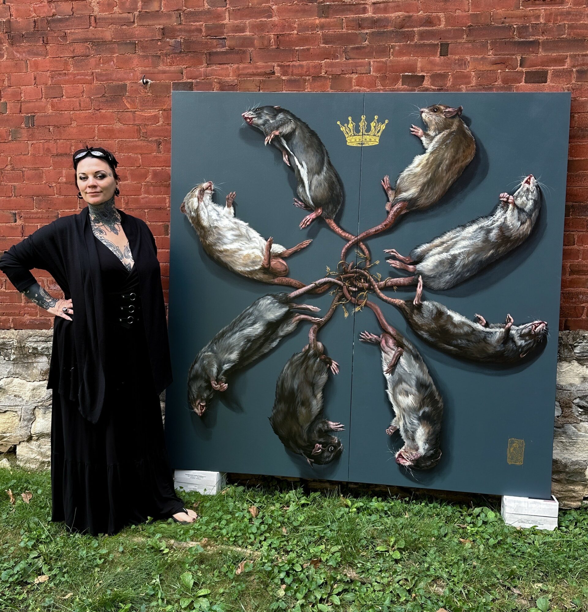Woman standing next to a large artwork of rats arranged in a circle with a crown above them, outdoors against a brick wall.