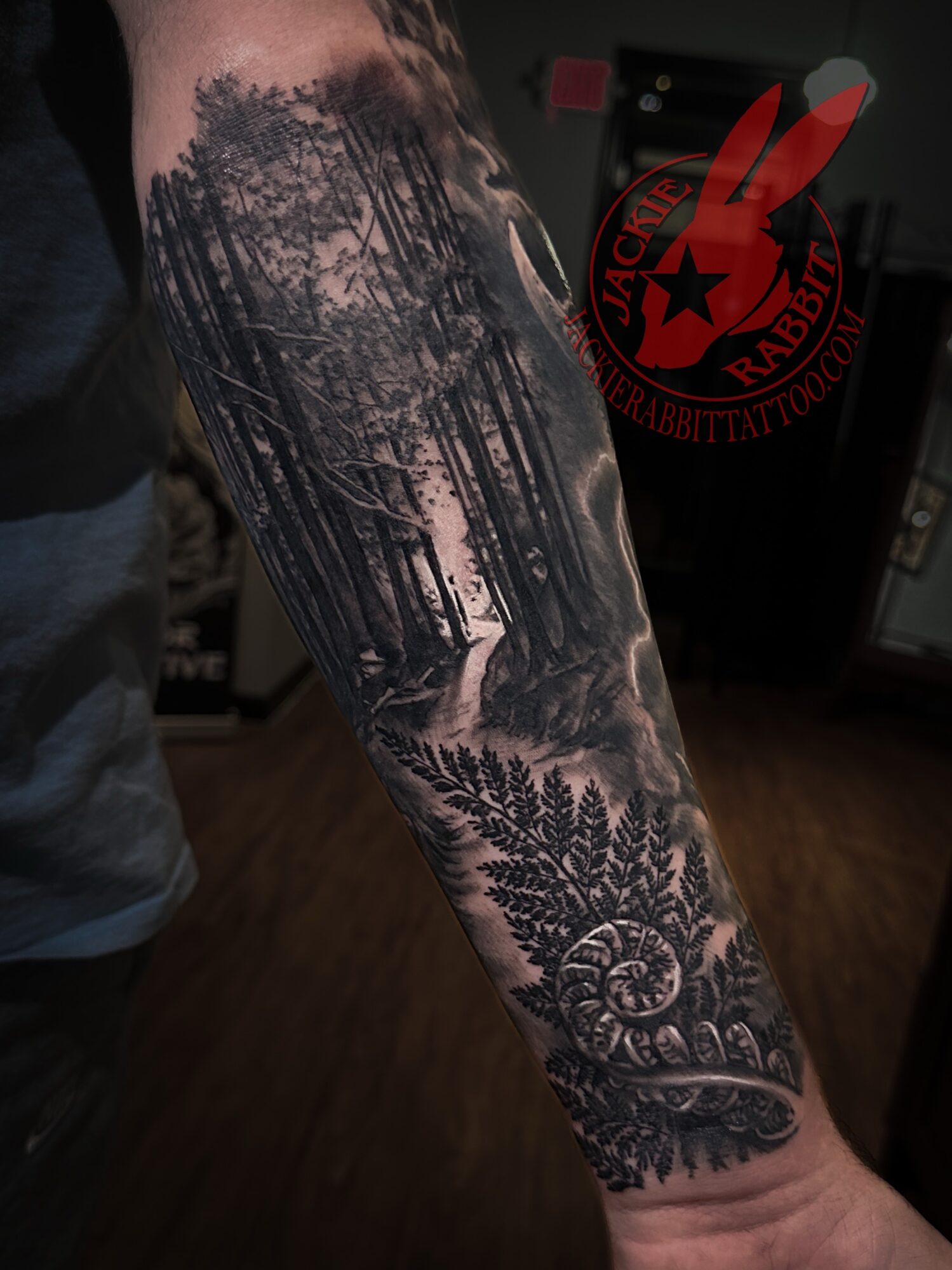 Forearm tattoo of a forest scene with trees, mountains, and detailed foliage, in black and gray ink.
