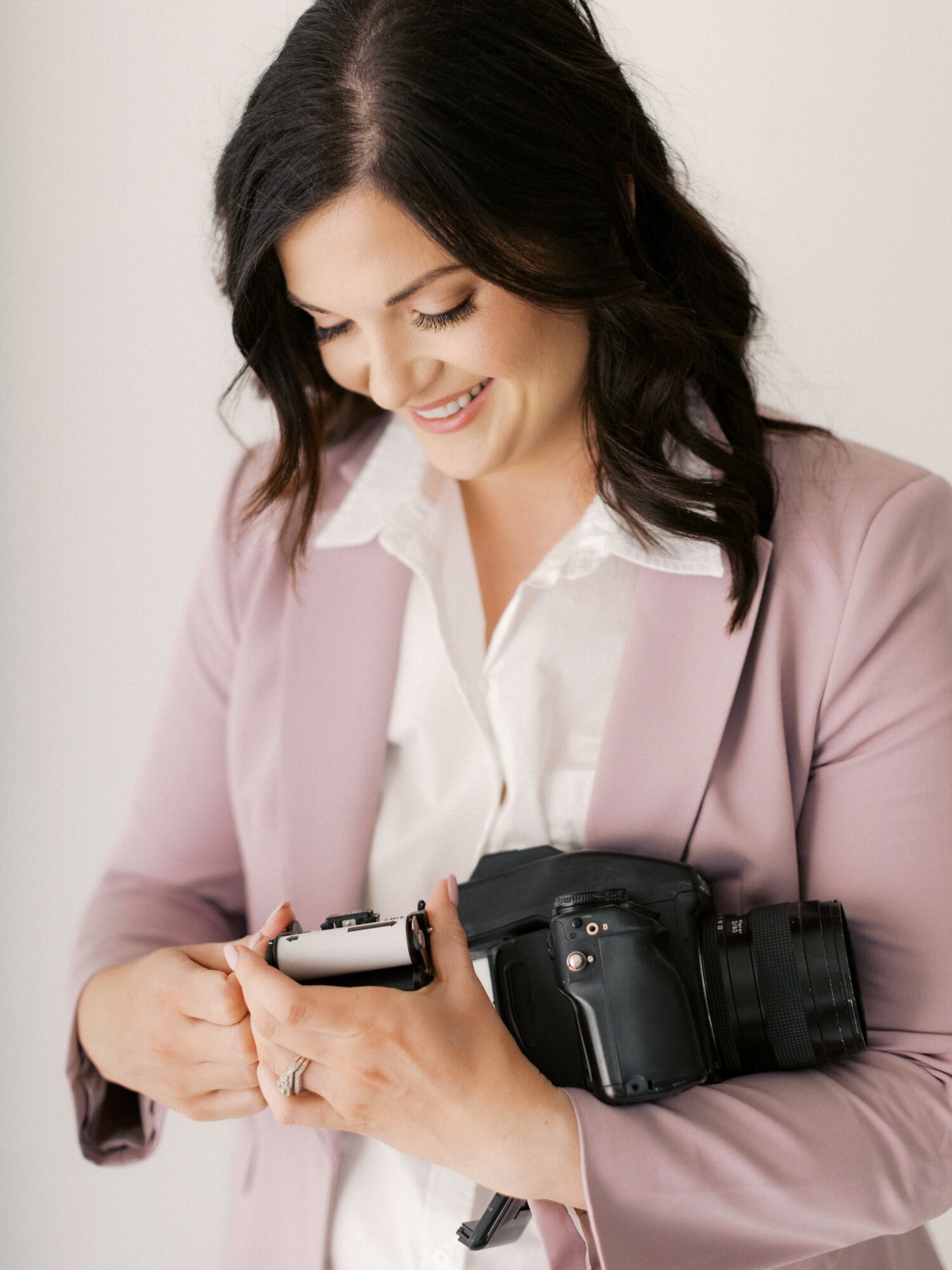 Woman with dark hair in a light pink blazer and white shirt holding a camera, smiling, against a plain background.