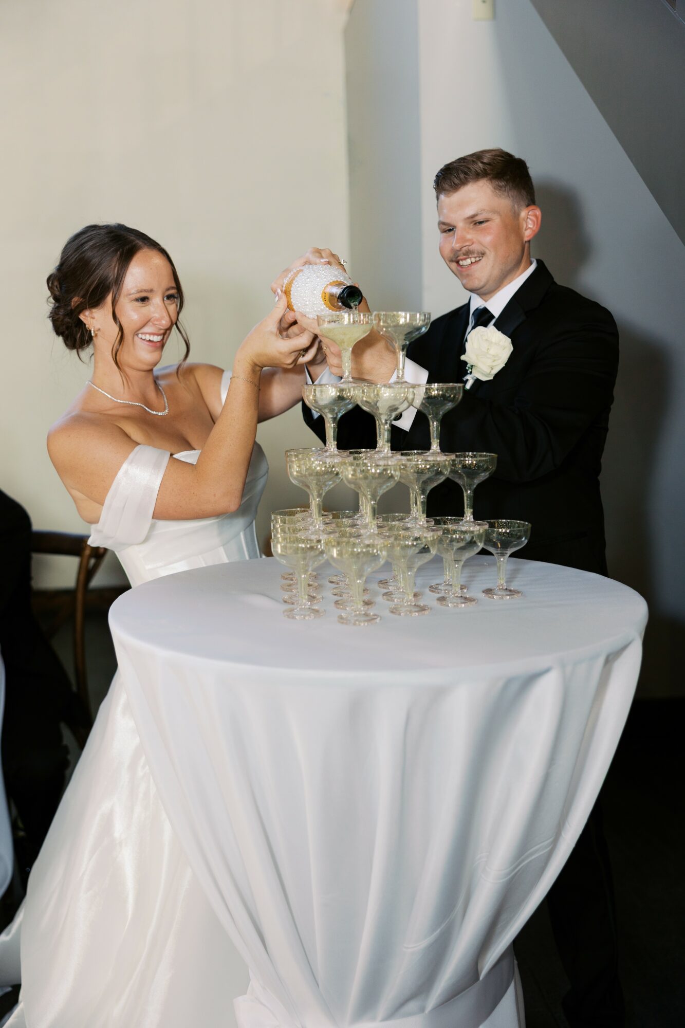 Bride and groom pouring champagne into a pyramid of glasses at a wedding reception, smiling, with a white tablecloth-covered table.
