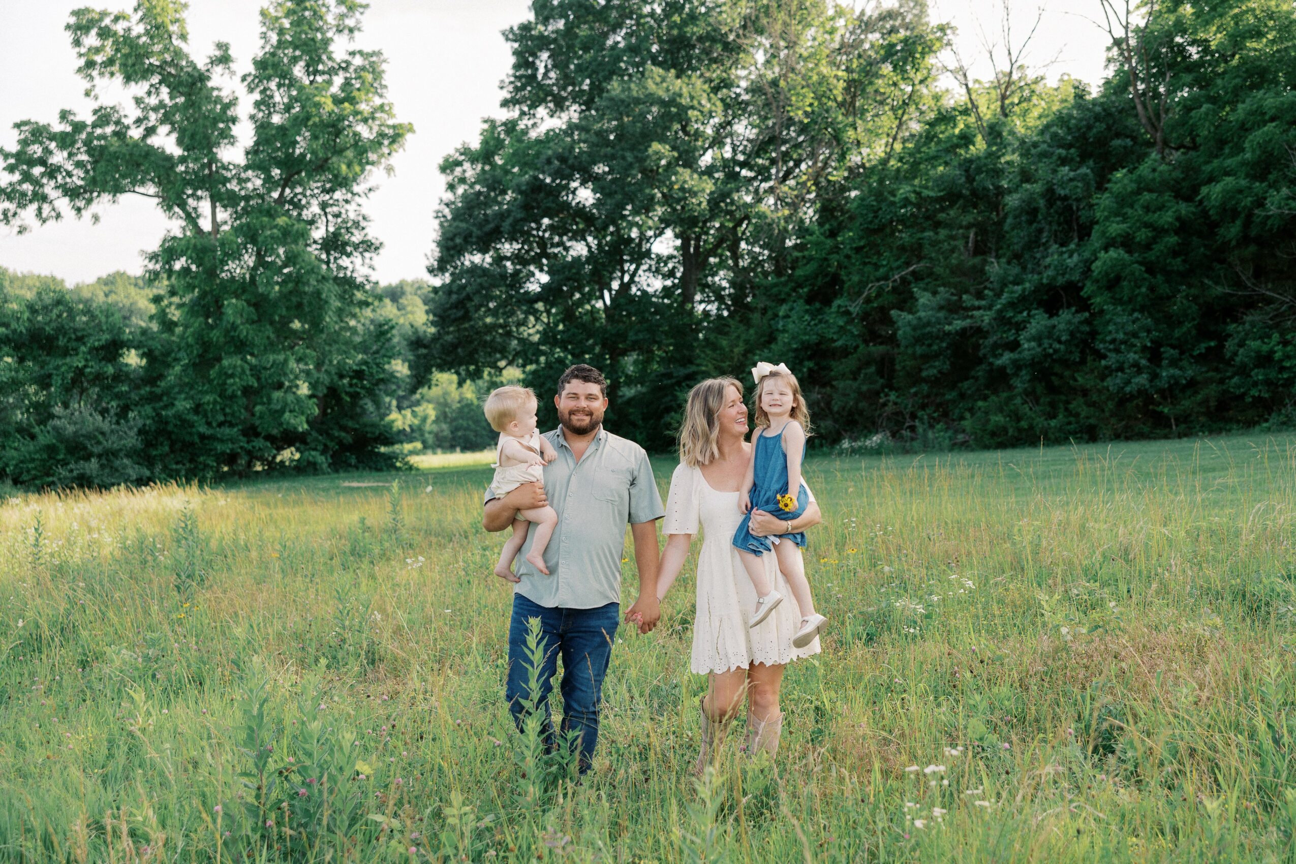 Family walking in a grassy field with trees in the background, holding children, during daytime.