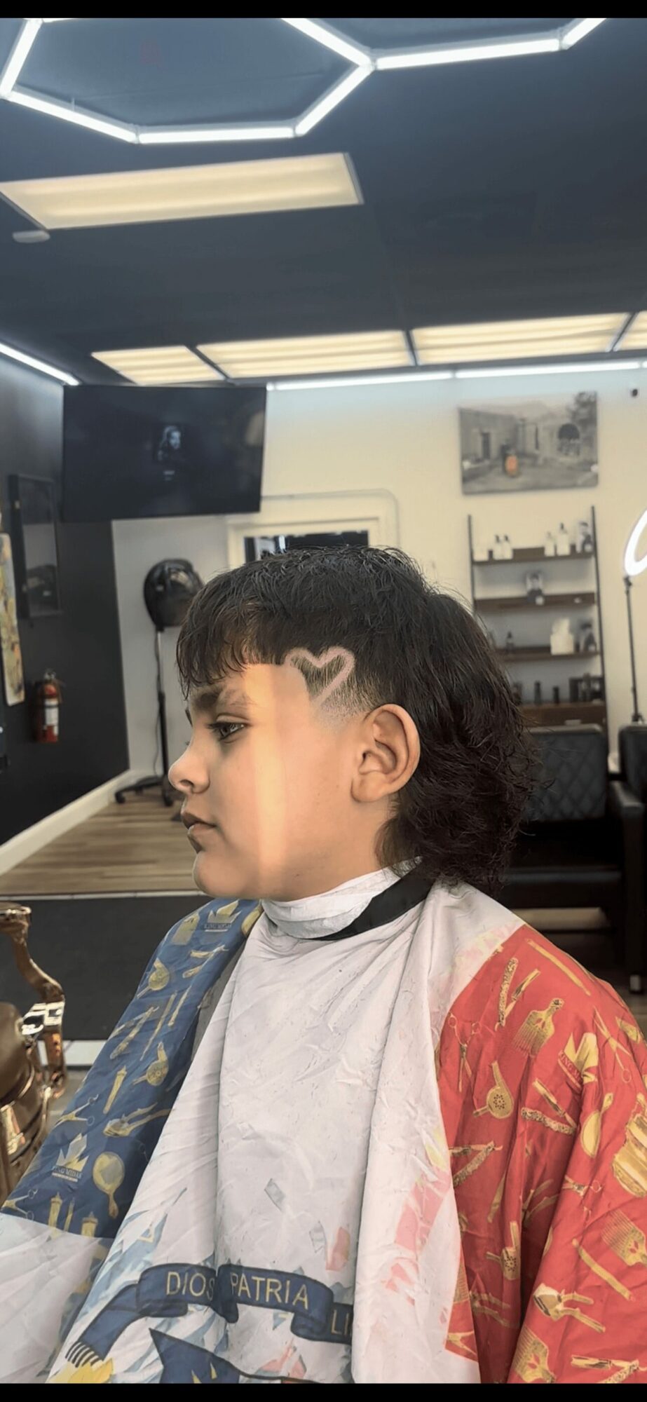 Person with short curly hair and a heart-shaped mark on the forehead, sitting in a salon with mirrors and lighting above.