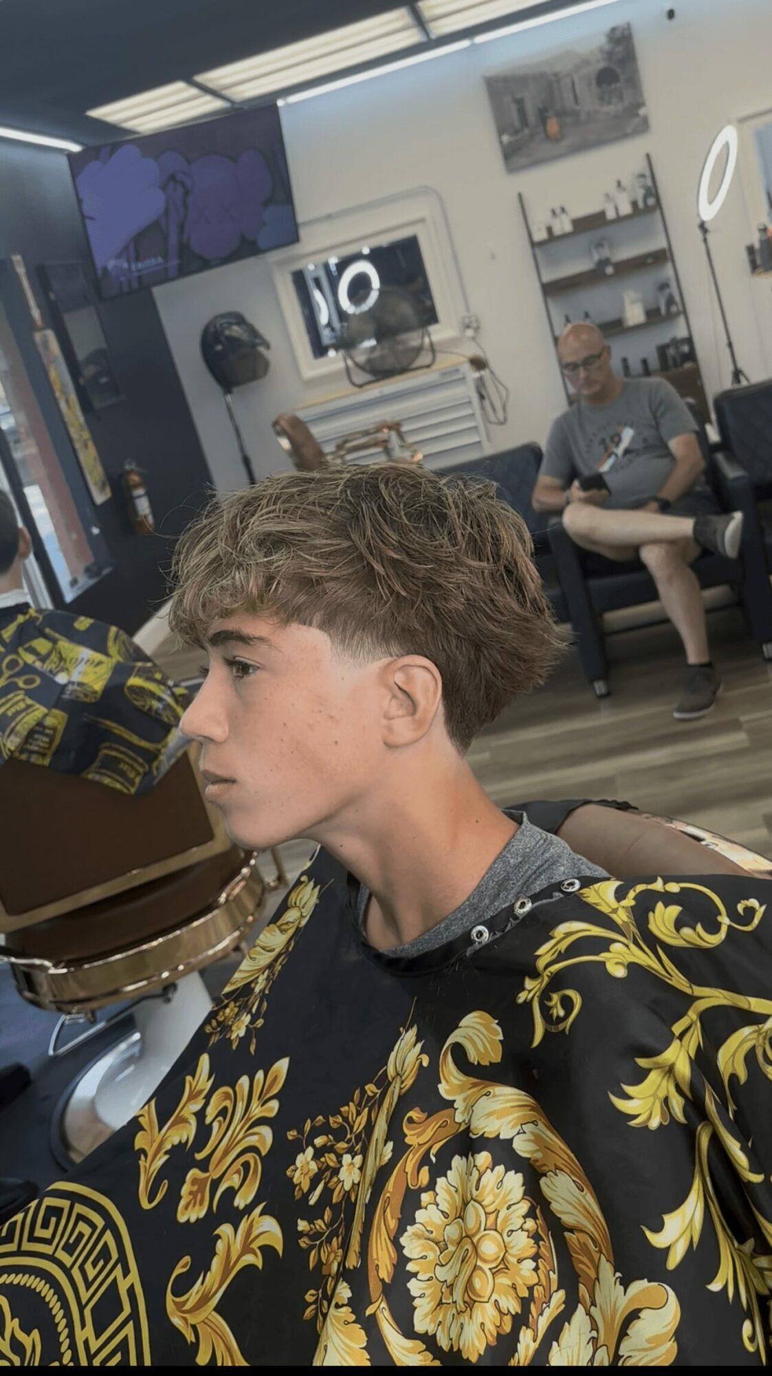 Young person with styled hair in a patterned shirt, sitting in a barbershop or salon, with mirrors and people in the background.