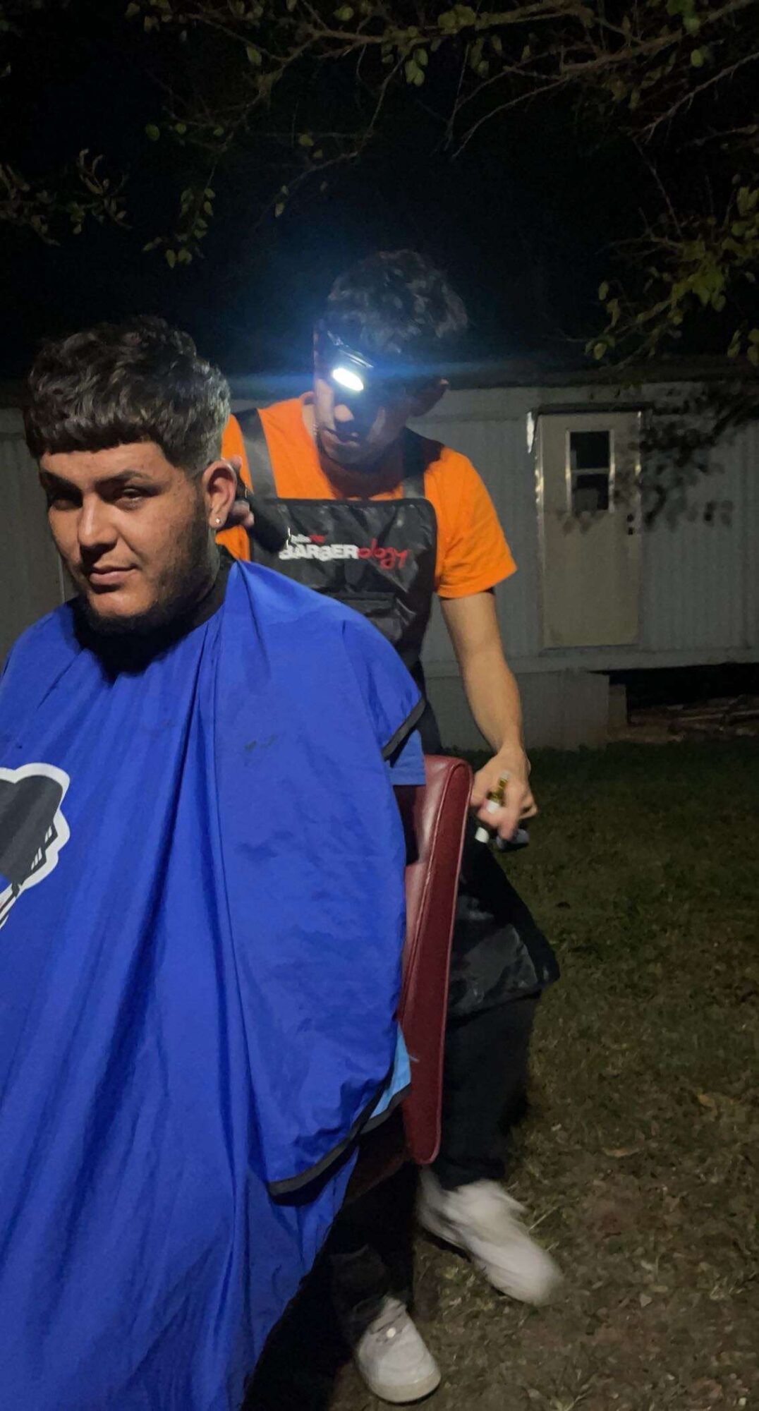 Person receiving a haircut outdoors at night, with another person standing behind wearing a headlamp, trees, and a utility box in background.