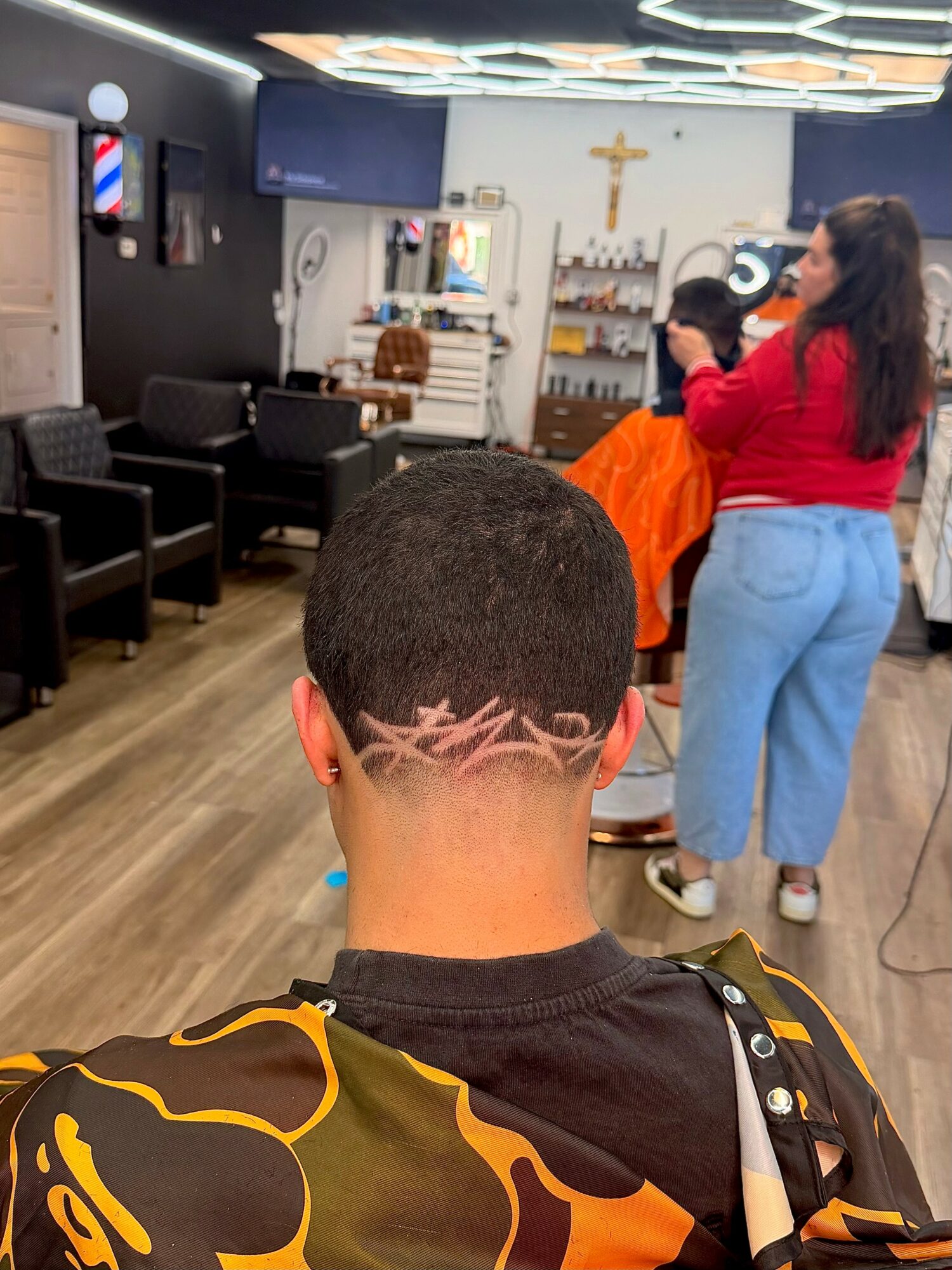 Person with a short haircut and a tattoo on the back of the neck, sitting in a salon or barbershop, with people and salon equipment in the background.