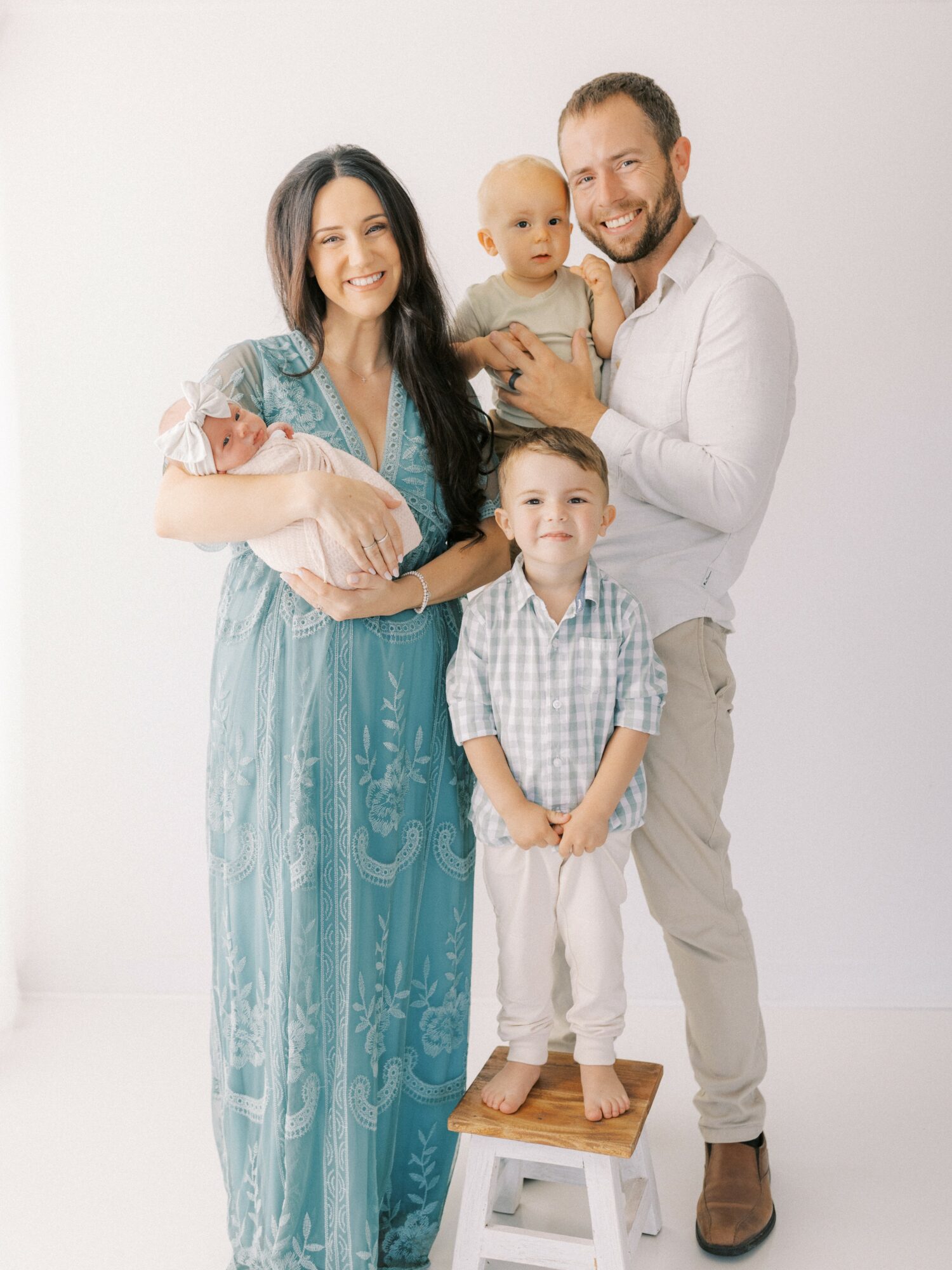 Family of five smiling, woman holding a baby, man holding a toddler, boy standing on a stool, all in casual clothing.