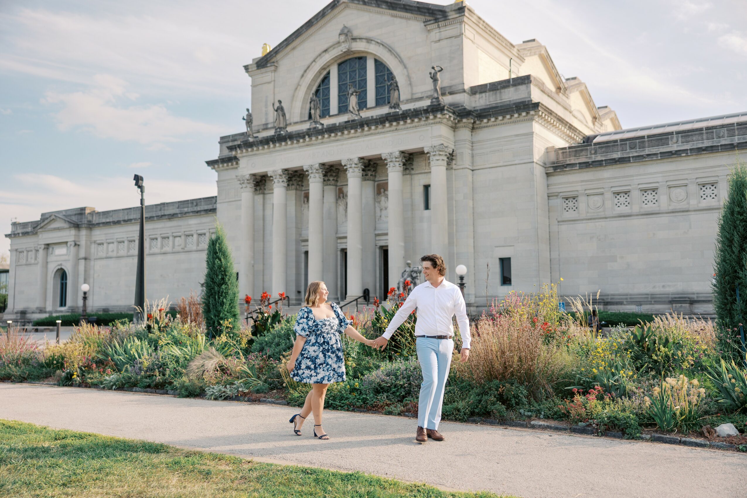 A couple holding hands walking in front of a large historic building with columns and a garden, daytime.