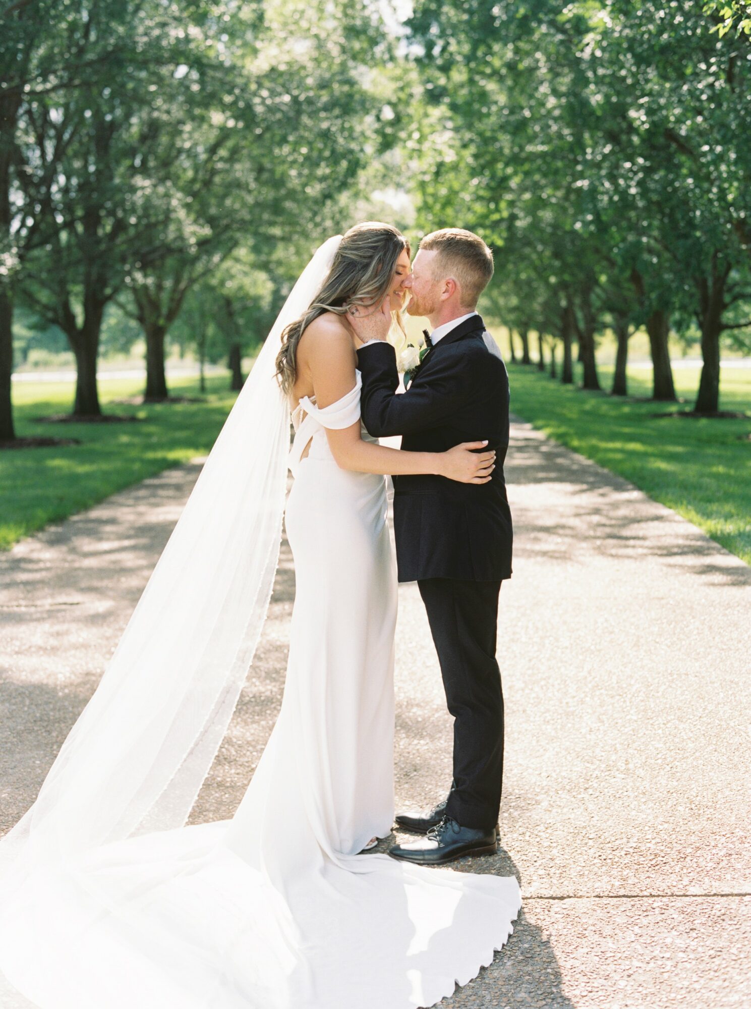 A bride and groom kiss on a tree-lined path outdoors, bride in a white gown with a long veil, groom in a black suit.
