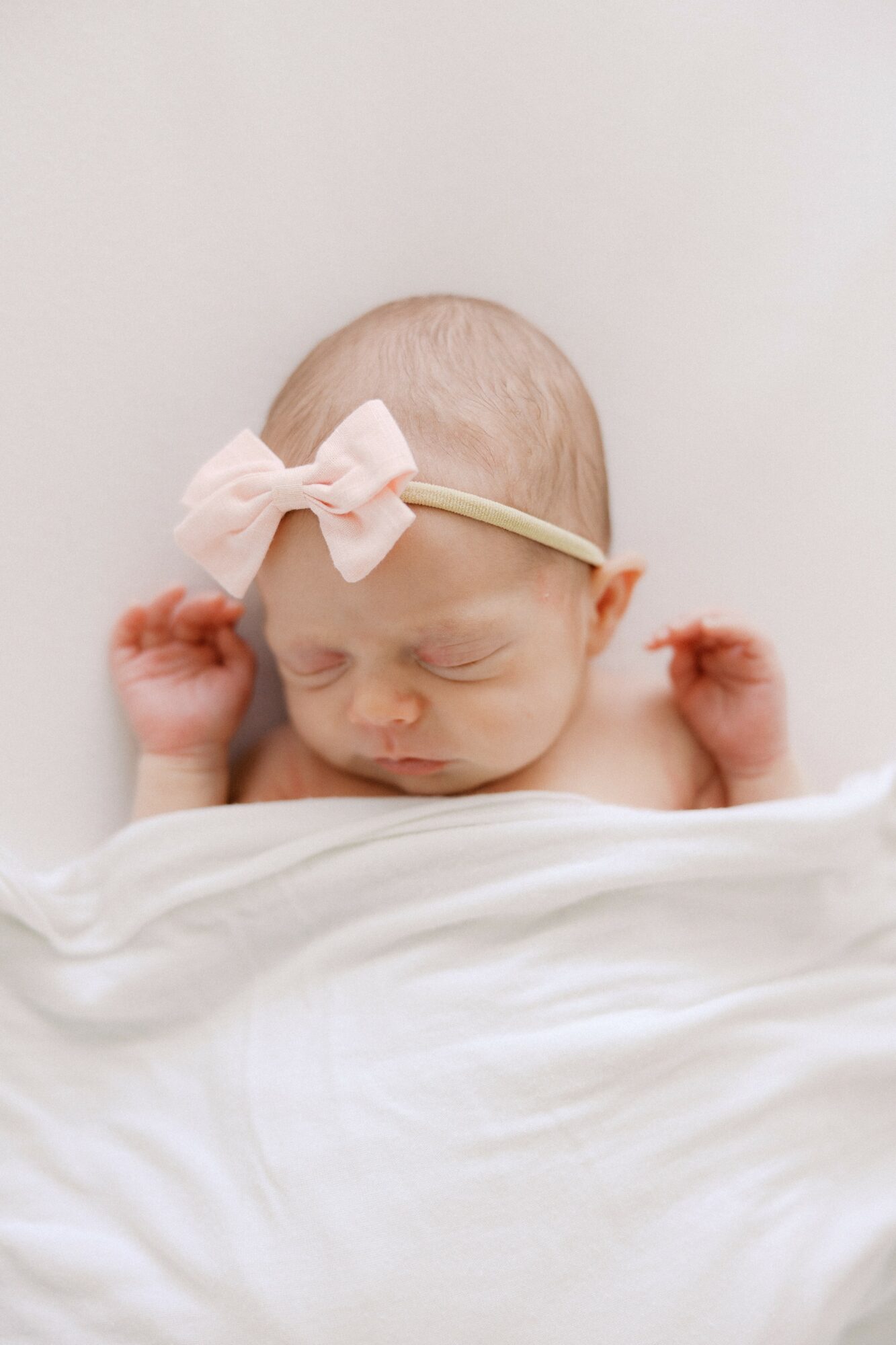 Newborn baby with a pink bow headband, sleeping with hands near face, wrapped in white blanket, against a plain background.