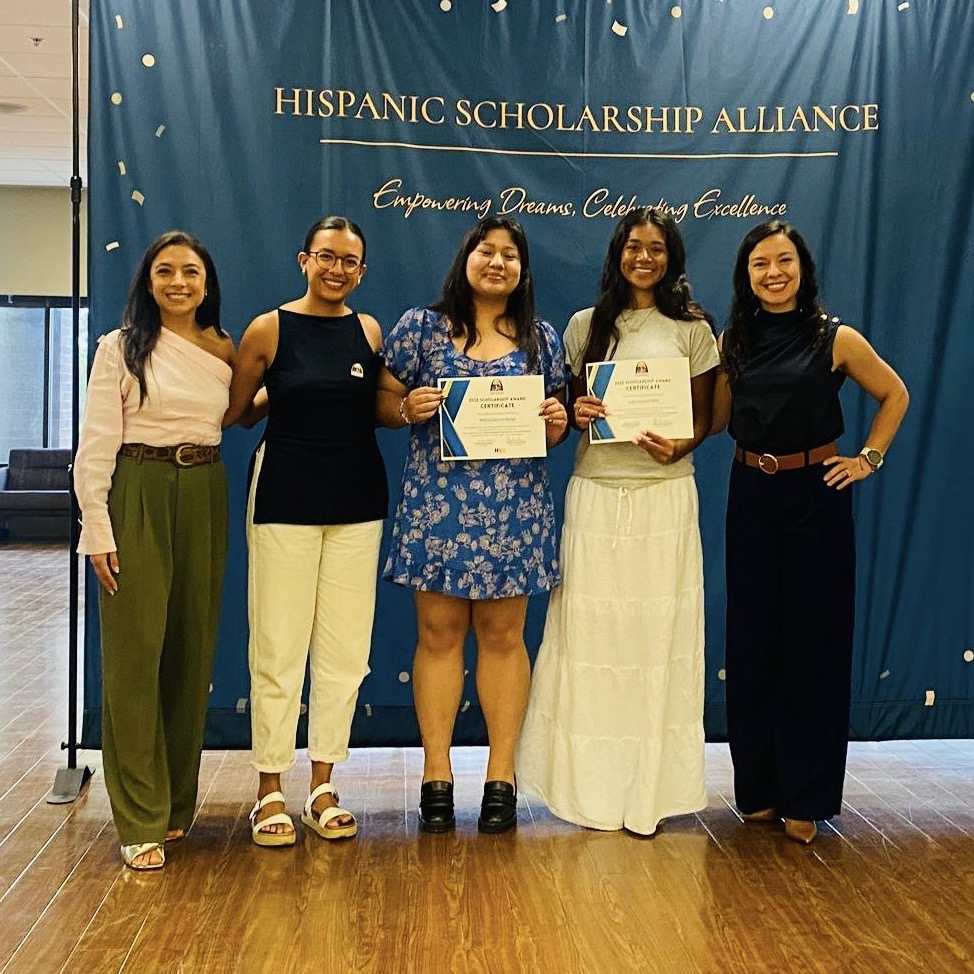 Five women standing on stage in front of a blue backdrop with gold text, holding certificates, smiling. Wood floor beneath them.