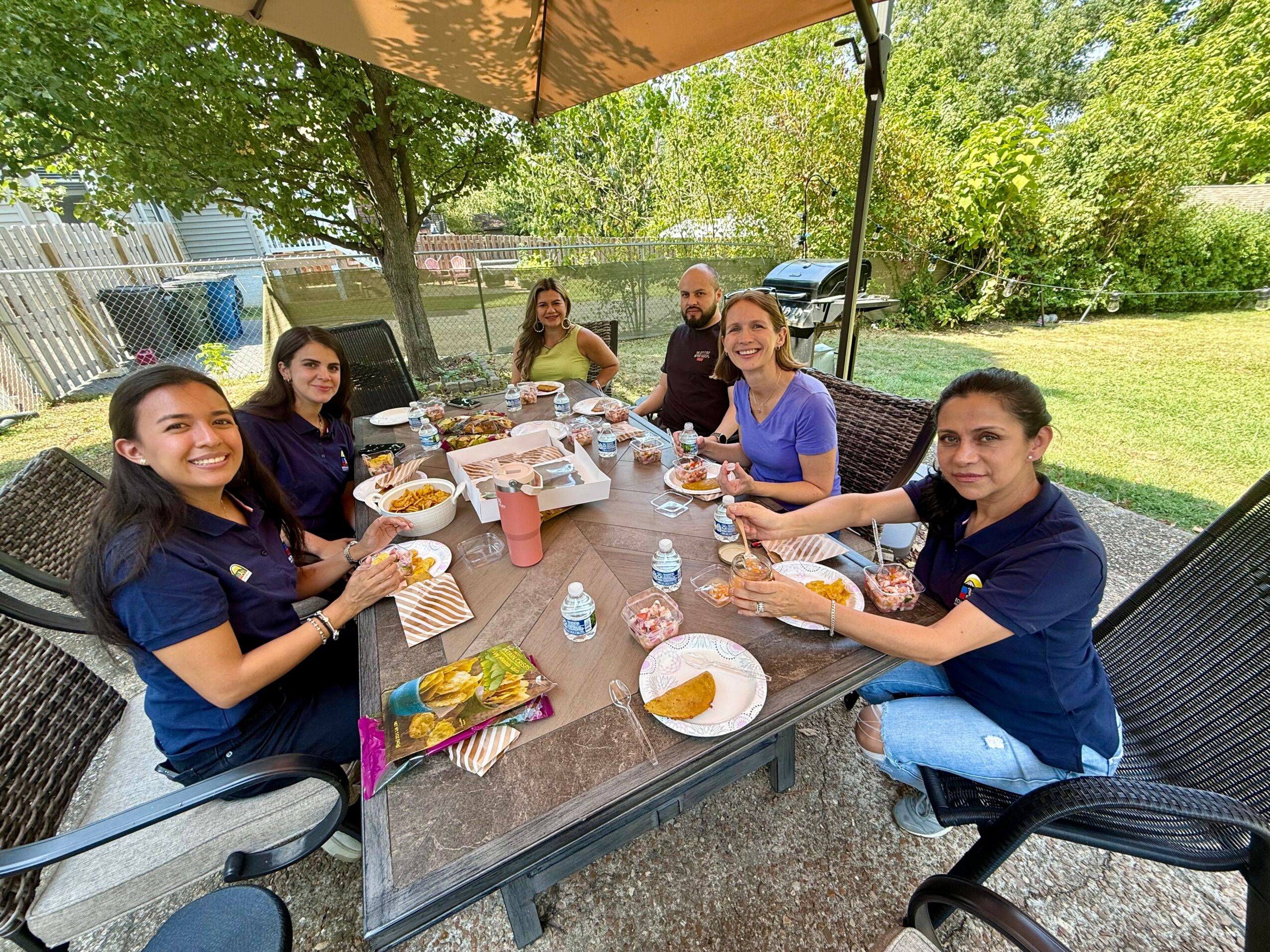 Group of seven people sitting around a table outdoors, enjoying food and drinks under a canopy, with trees and cars in background.