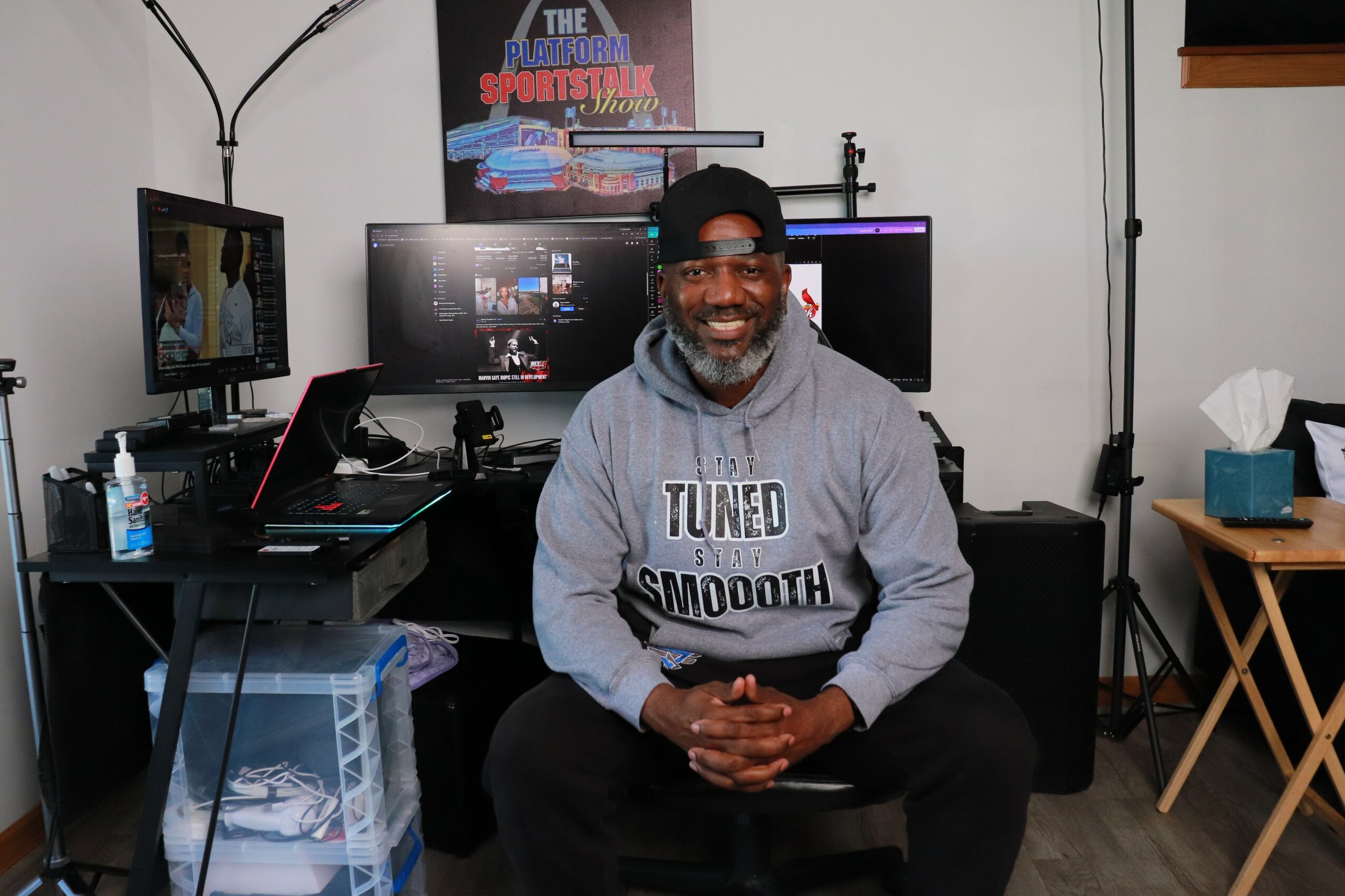 Smiling man with glasses and a beard sitting in front of multiple computer monitors in a room.