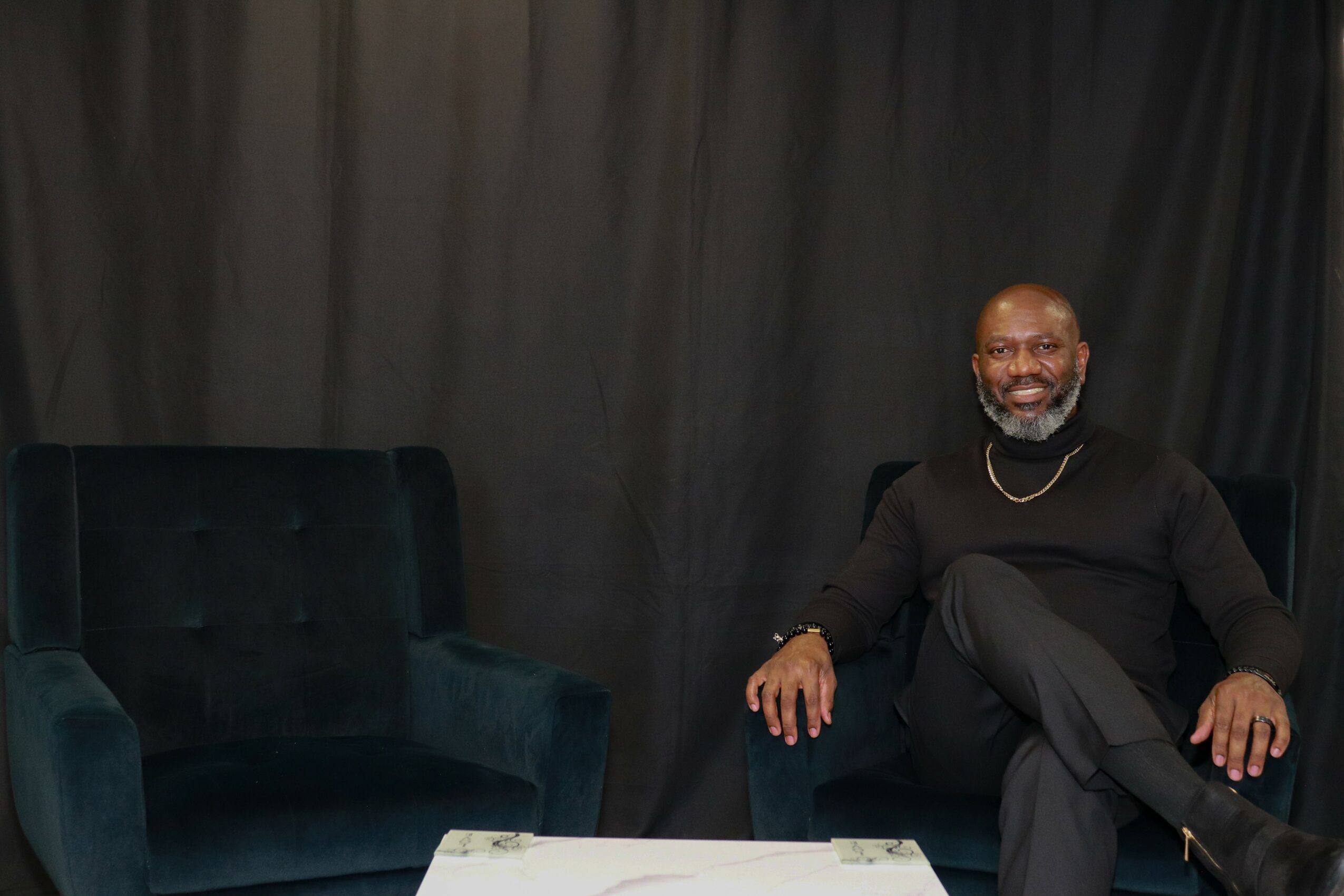 A smiling man with a beard and necklace sits on a dark blue sofa, with another similar sofa beside him, against a black curtain backdrop.