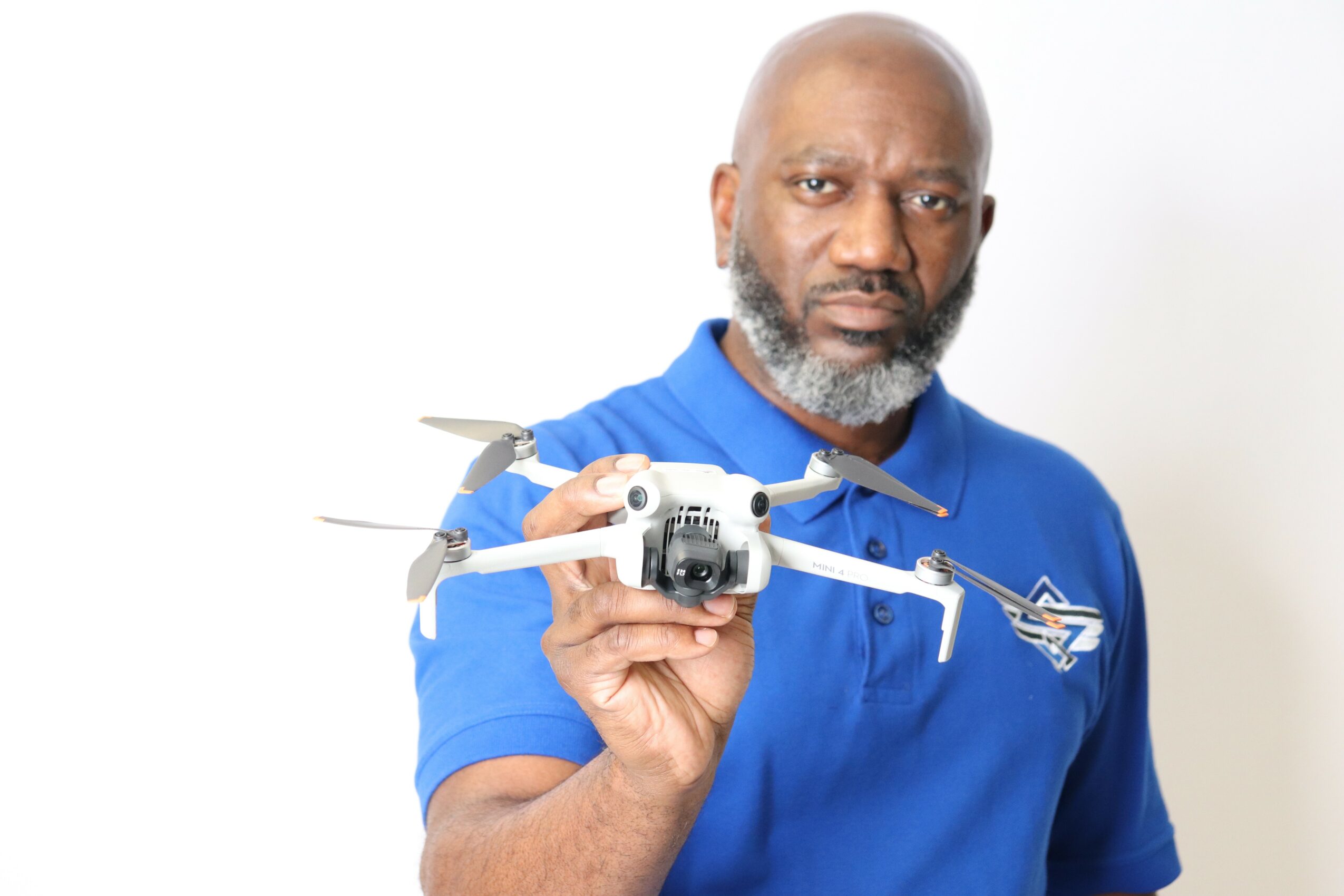 Man with a beard holding a drone, wearing a blue polo shirt with a logo, standing against a white background.