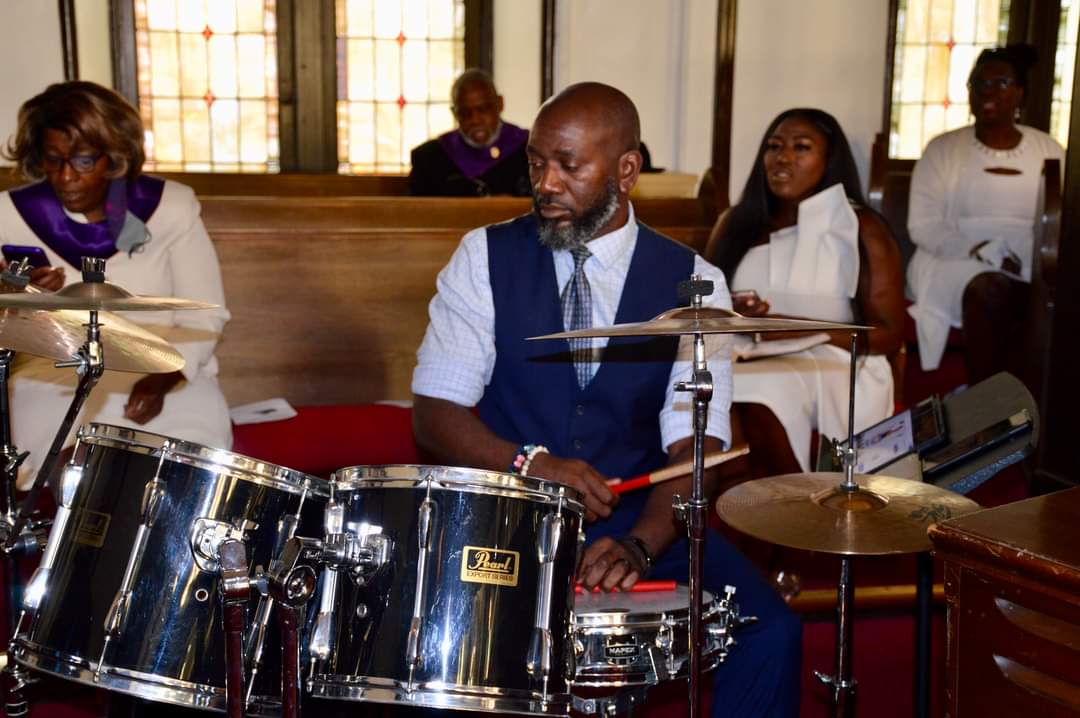 Man playing drums in a church with people seated in pews behind him.