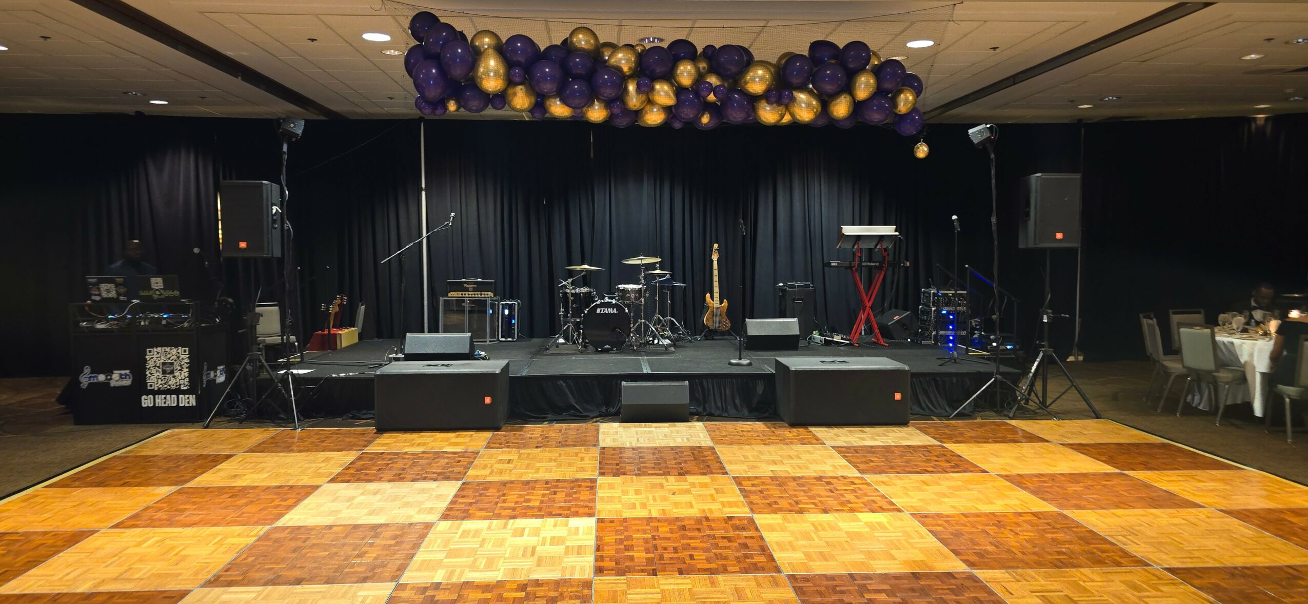 Stage with musical instruments and purple and yellow balloons overhead, in a dimly lit room with a checkered dance floor.