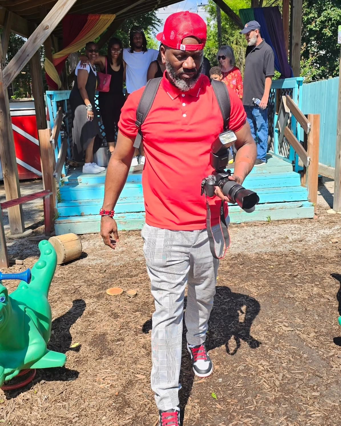 Man in red shirt and gray pants holding a camera, walking outdoors with a group in the background.