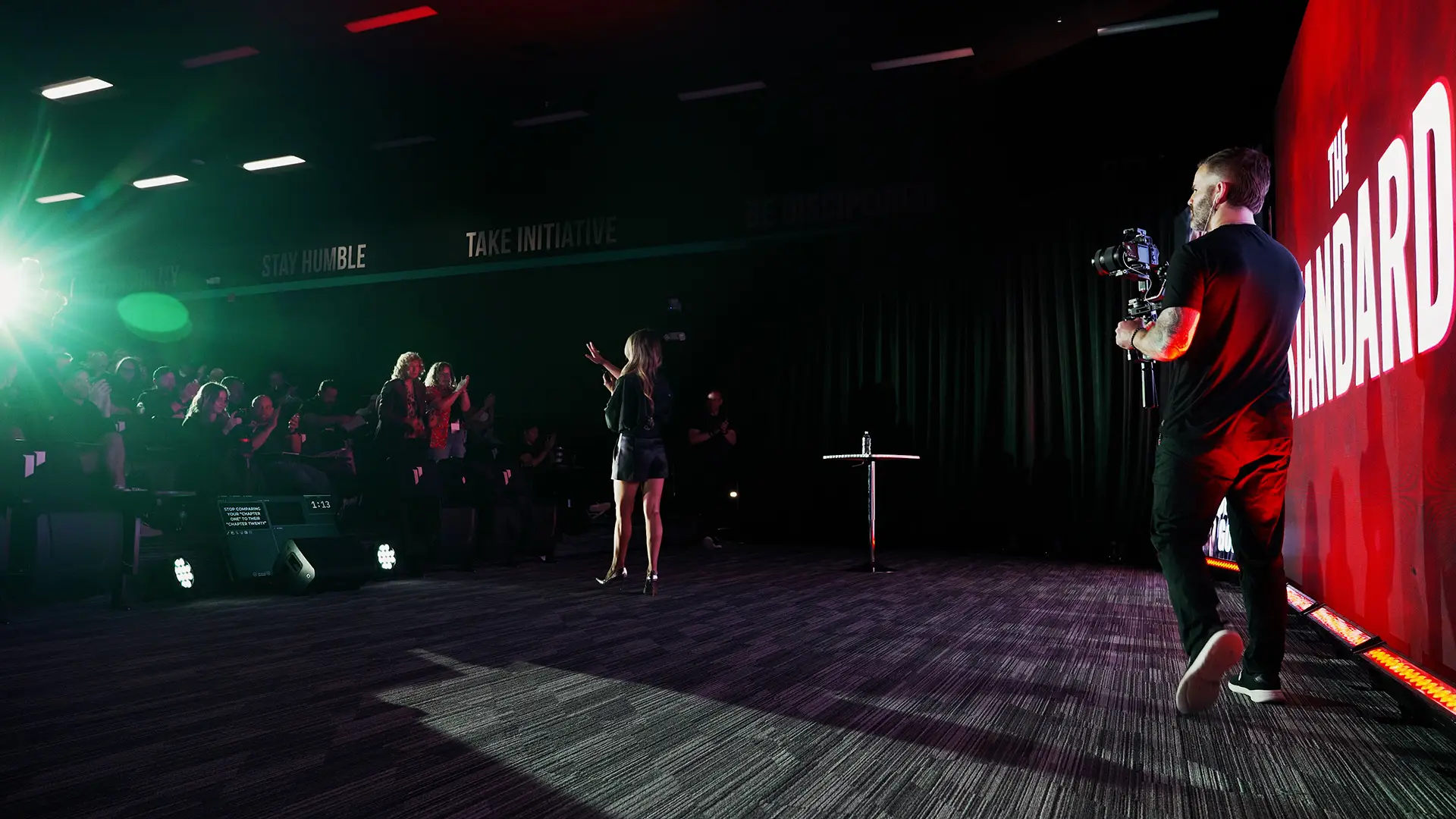 Person on stage speaking to audience with camera operator nearby, audience seated in dark room, bright stage lighting, red backdrop.
