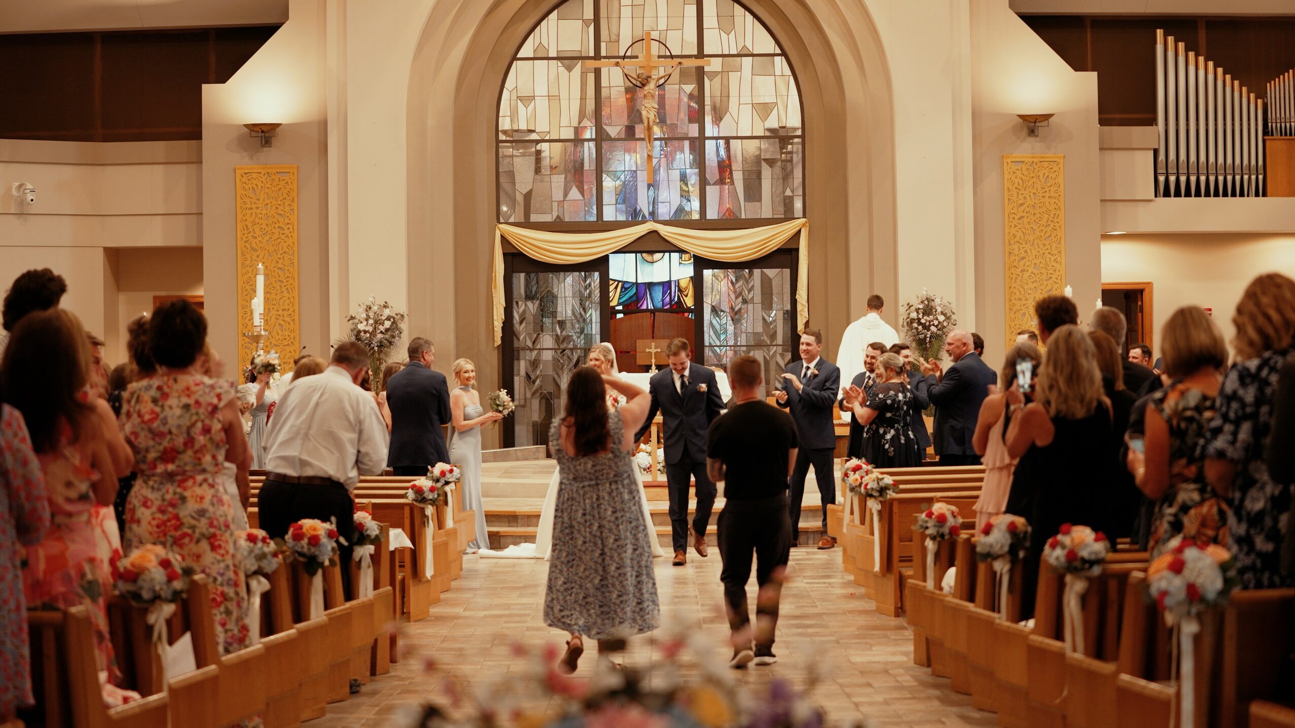 People walking down aisle toward altar in church with stained glass window, floral decorations, and congregation seated on sides.