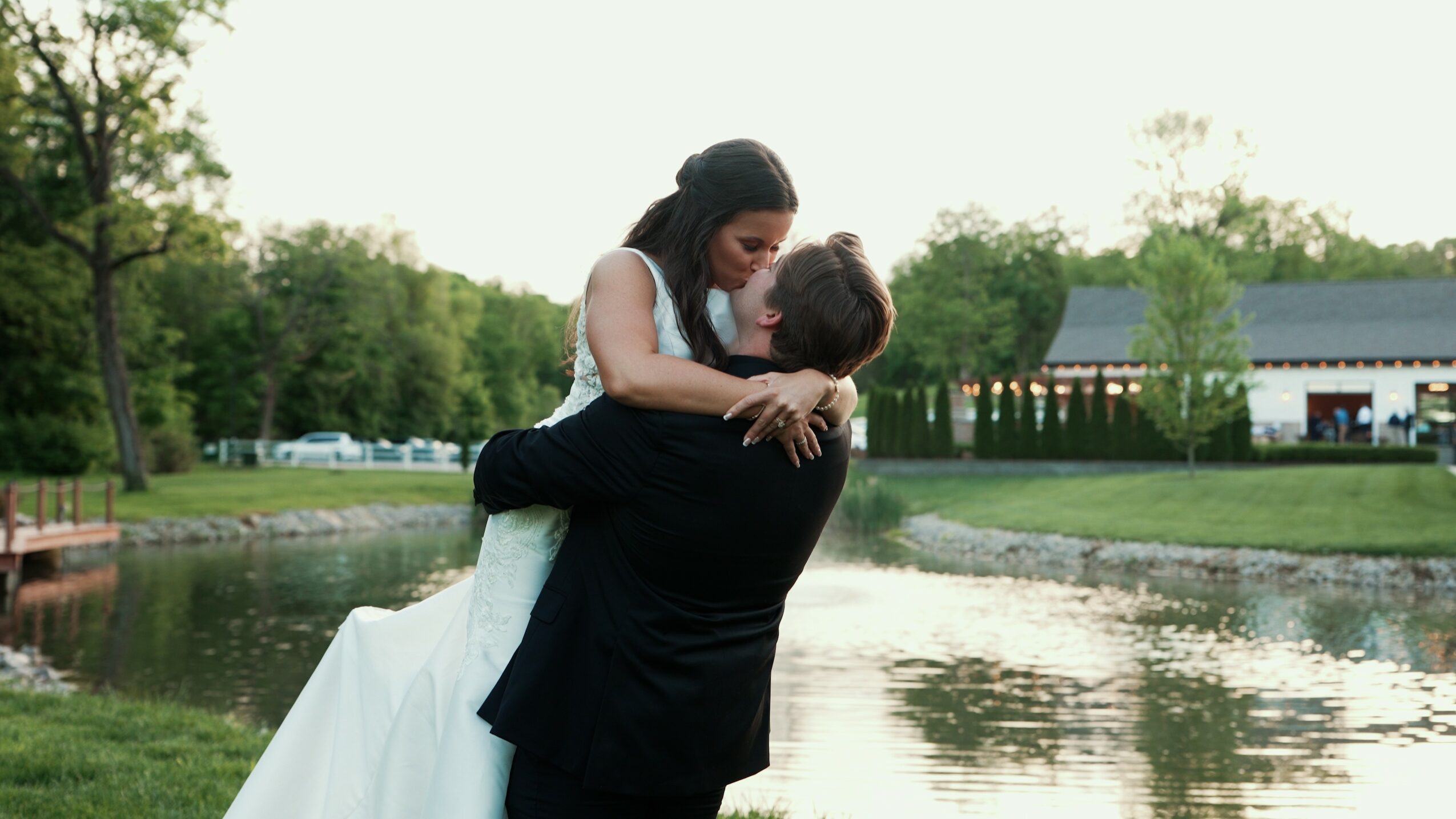 A woman in a wedding dress is lifted by a man near a pond outdoors, with trees and buildings in the background.