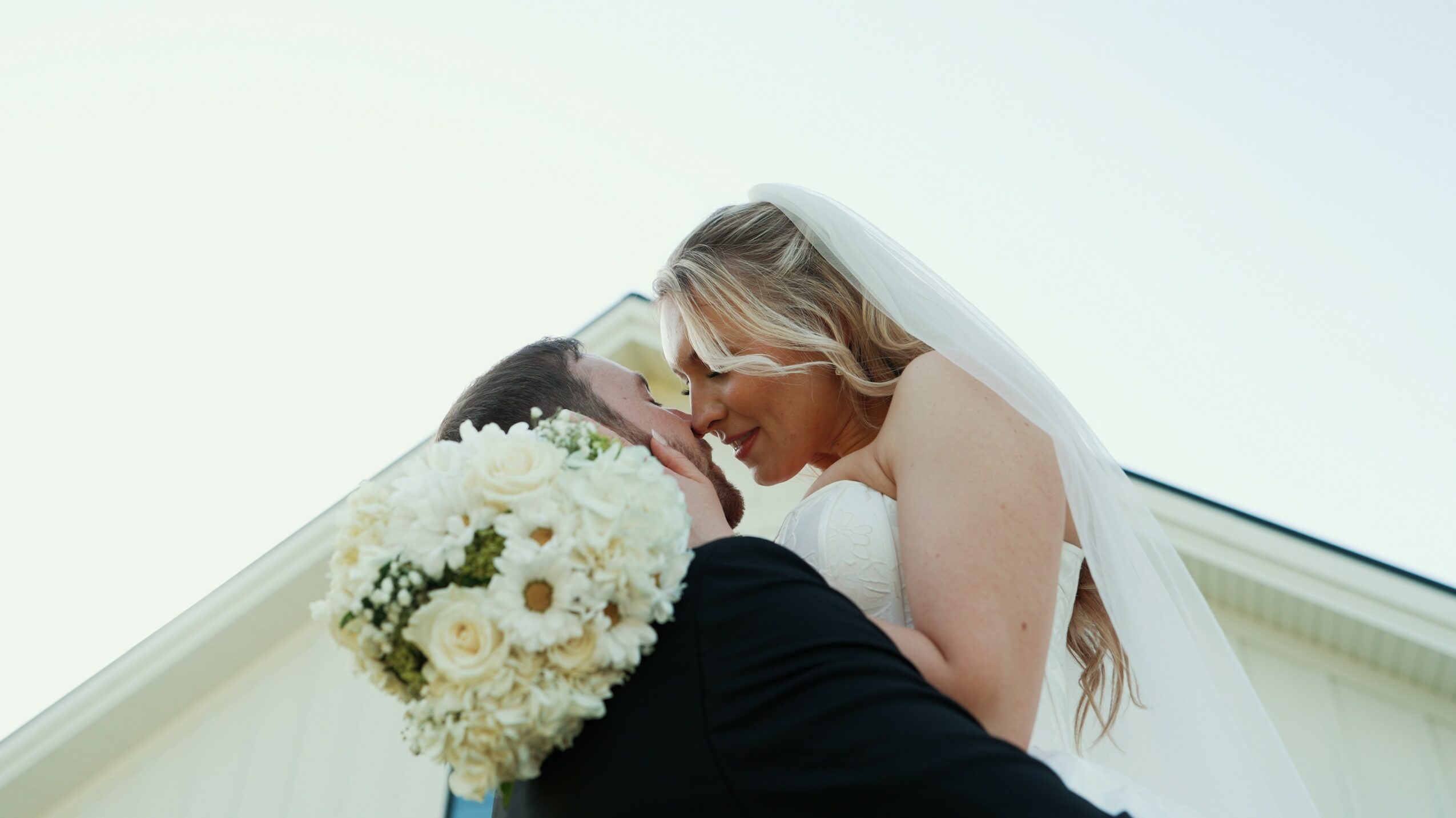 Bride and groom embrace, bride holding a large bouquet of white flowers, against a bright sky background.