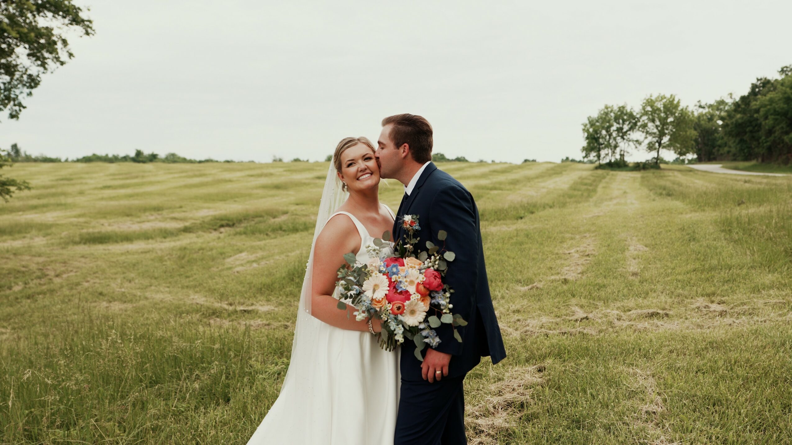 A bride and groom stand outdoors on a grassy field, smiling and holding a bouquet of flowers.