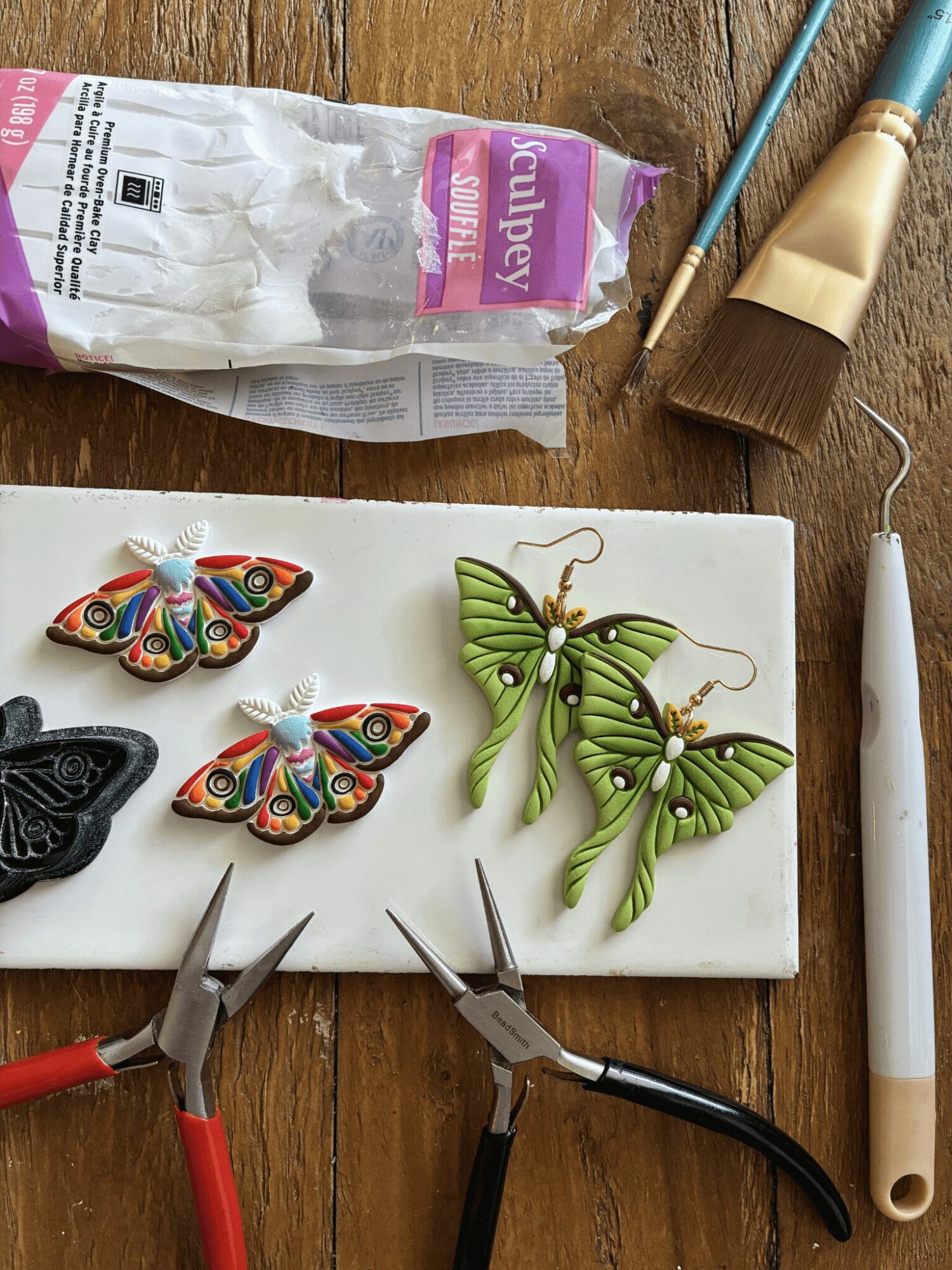 Colorful butterfly and green leaf earrings on a white surface with tools and supplies nearby.