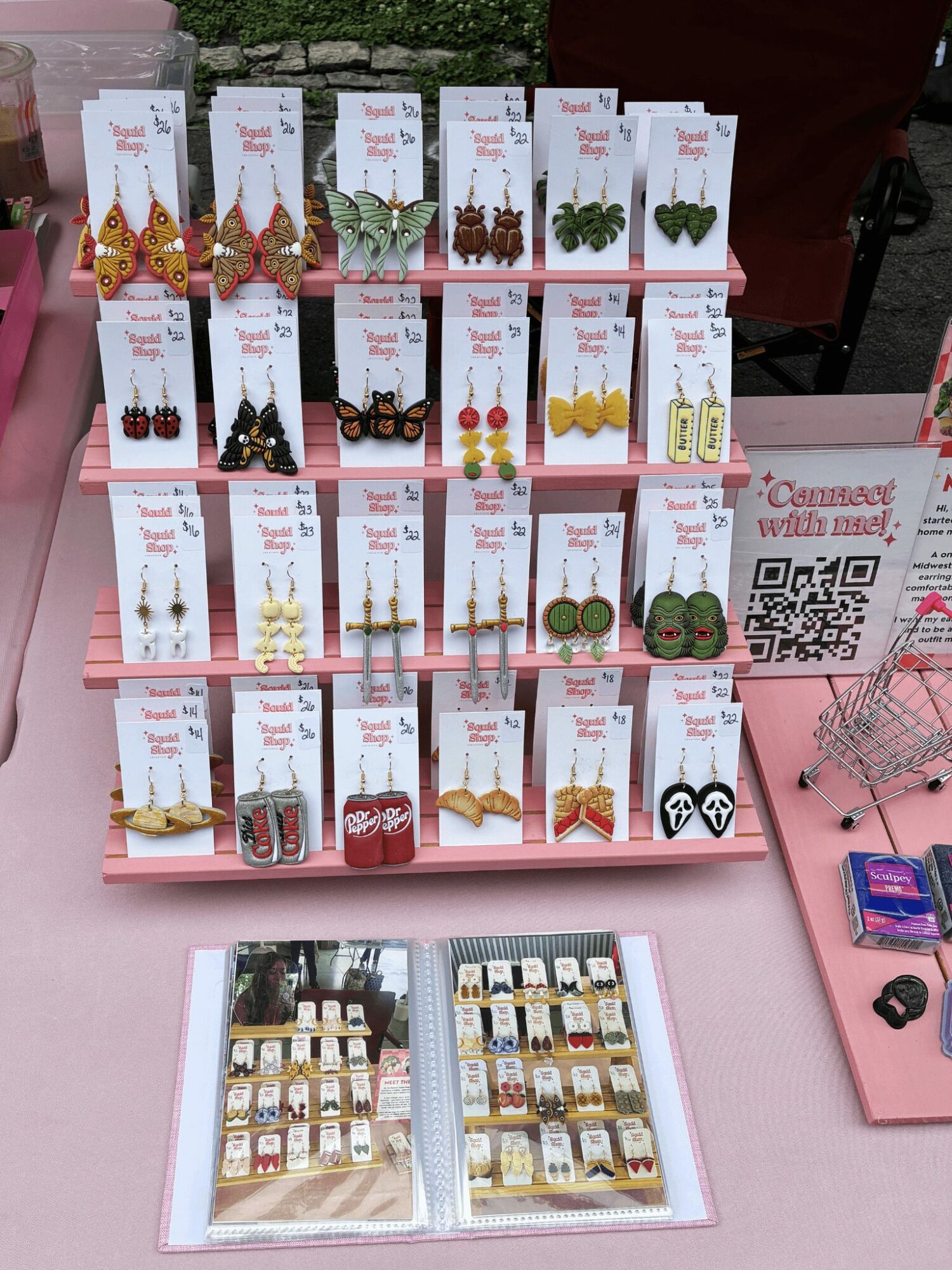 Display of various earrings on cards, arranged on a pink table with a QR code and small shopping cart, in an outdoor setting.