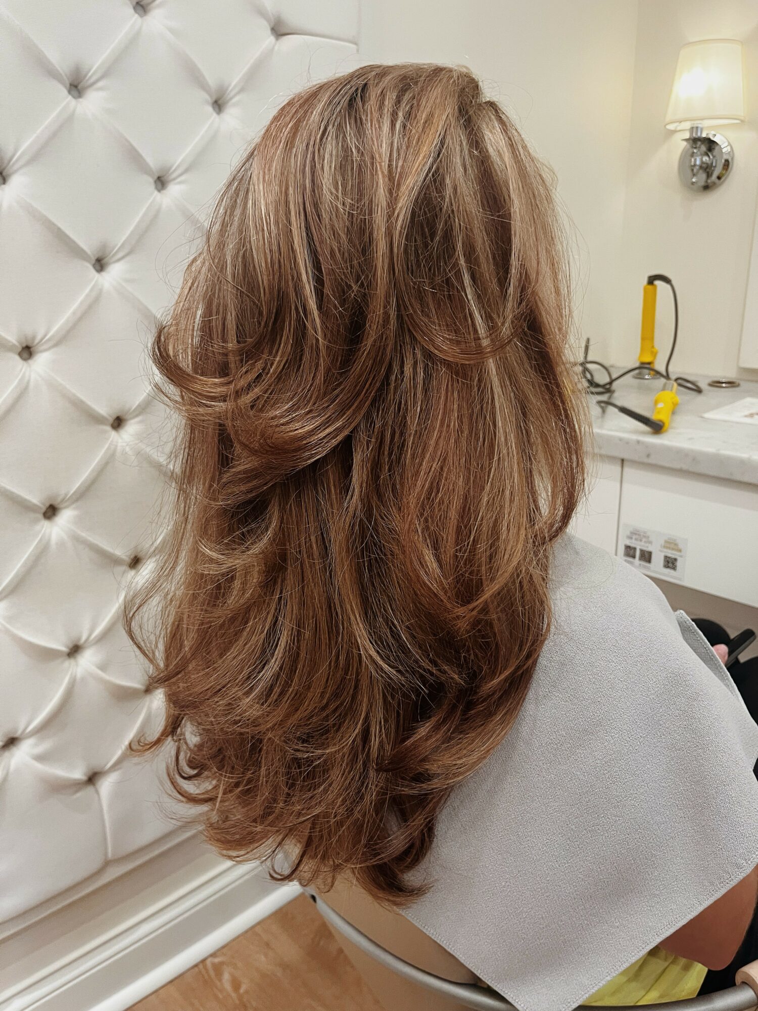 Side view of a woman with long, wavy, light brown hair in a salon setting, with a padded wall and lamp in background.