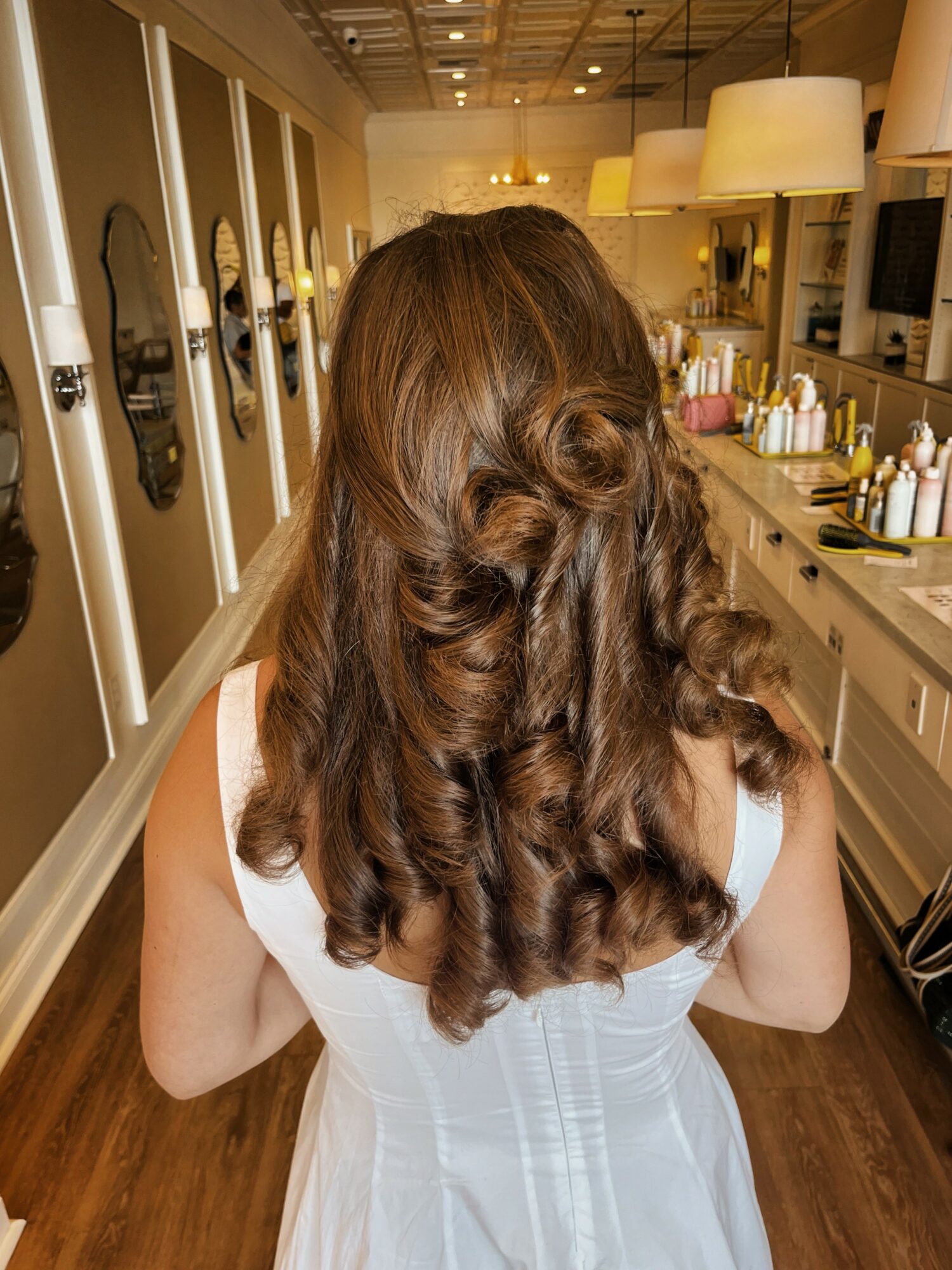 Back view of a woman with long, curly brown hair in a salon, wearing a white dress, with salon mirrors and products visible.