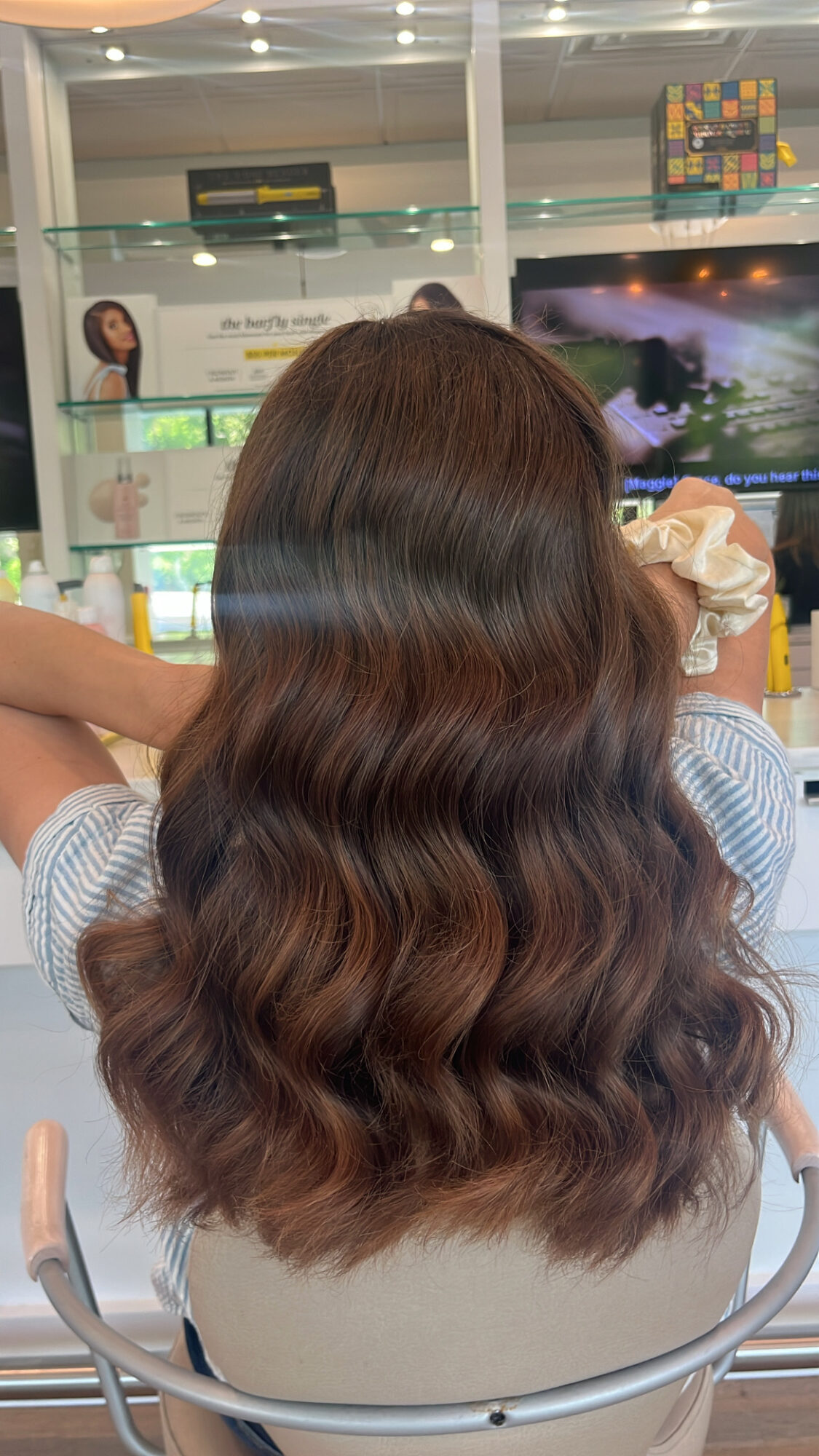 Back of a person with long, wavy brown hair sitting in front of a television screen in a store or public space.