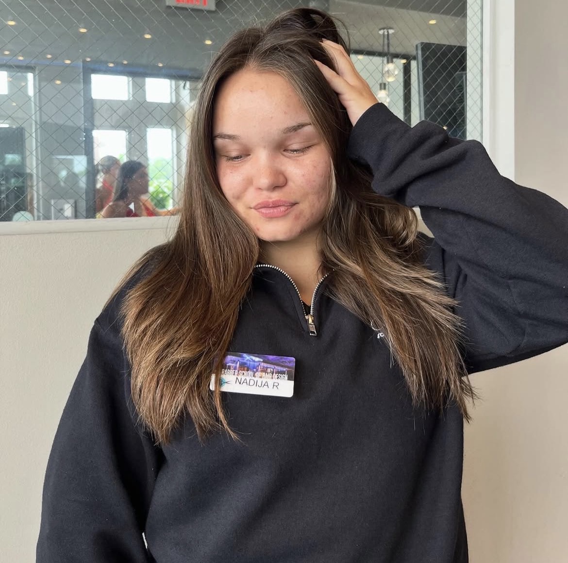 Young woman with long brown hair, eyes closed, hand on head, wearing a black zip-up top, indoors.