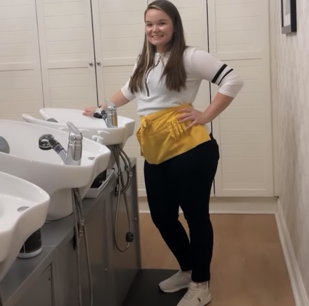 Young woman standing in a bathroom with sinks, smiling, wearing a white top, yellow apron, black pants, and white shoes.