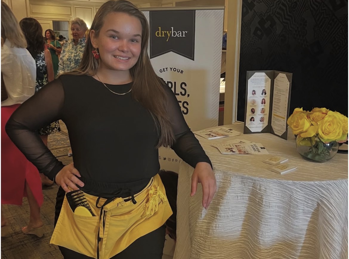 Young woman with long hair smiling, standing next to a table with yellow flowers and promotional materials.