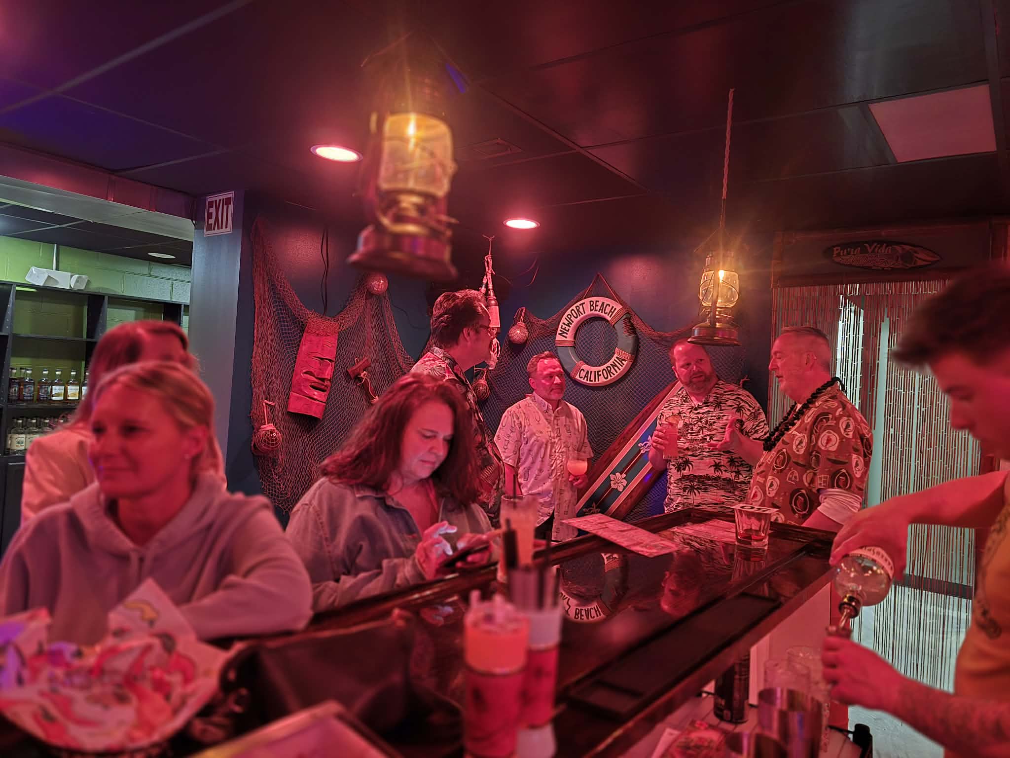 People gather at a bar with red lighting, some using phones, others talking, and a bartender serving drinks.