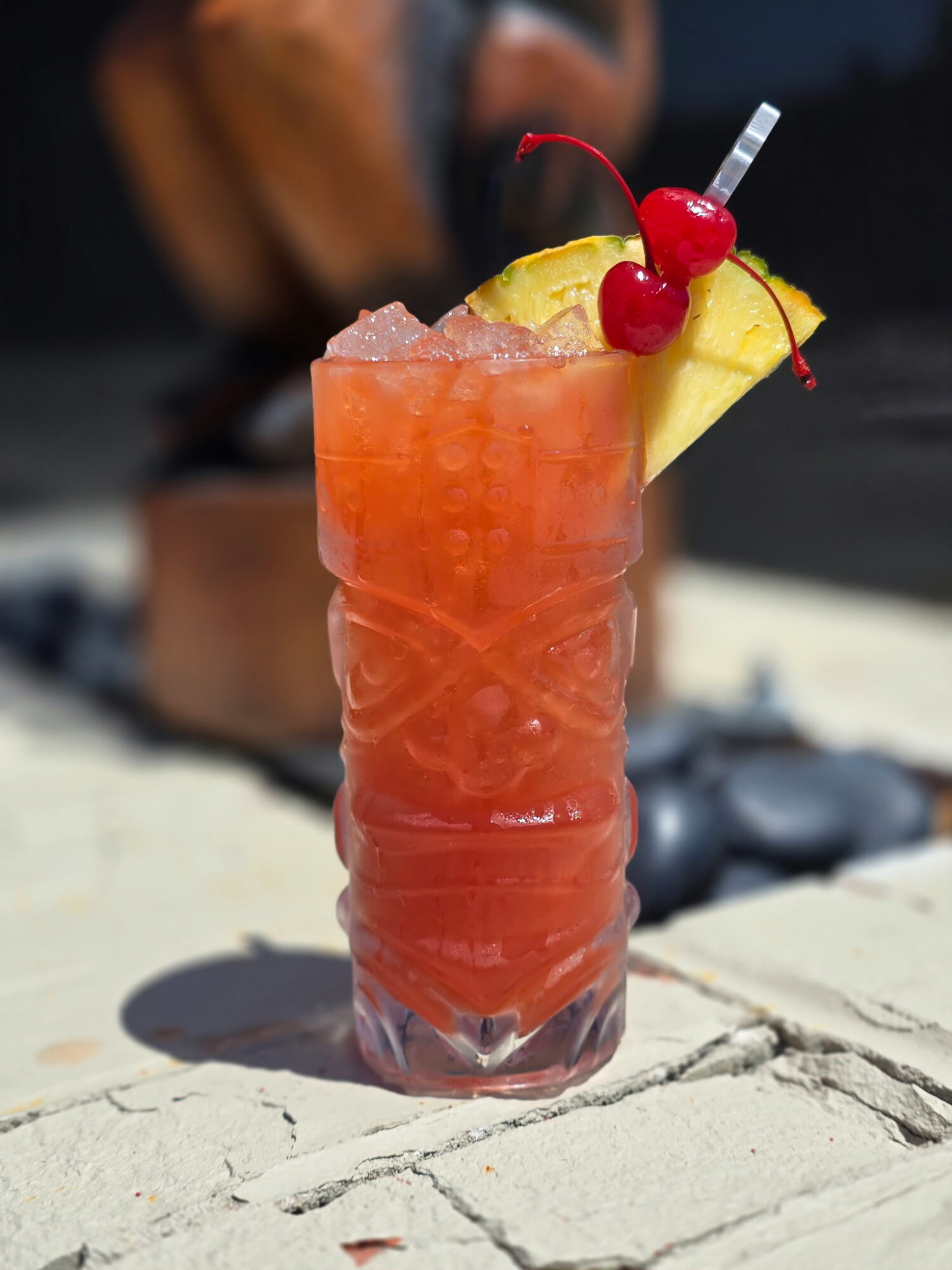 Tall glass of pink cocktail with pineapple slice and cherries, on a light-colored surface outdoors.