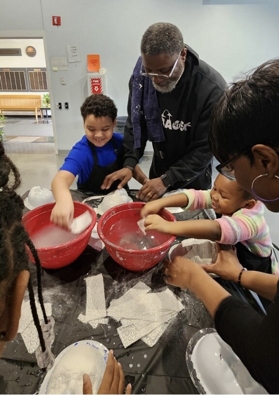 Adult and children mixing ingredients in red bowls at a table, with a light blue wall in the background.