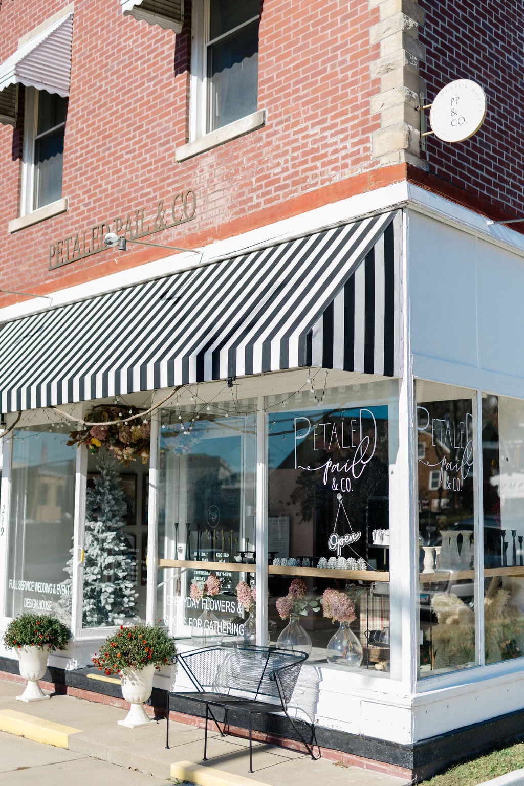 Corner of a brick building with a striped awning over a glass storefront, potted plants outside, and a bench nearby.