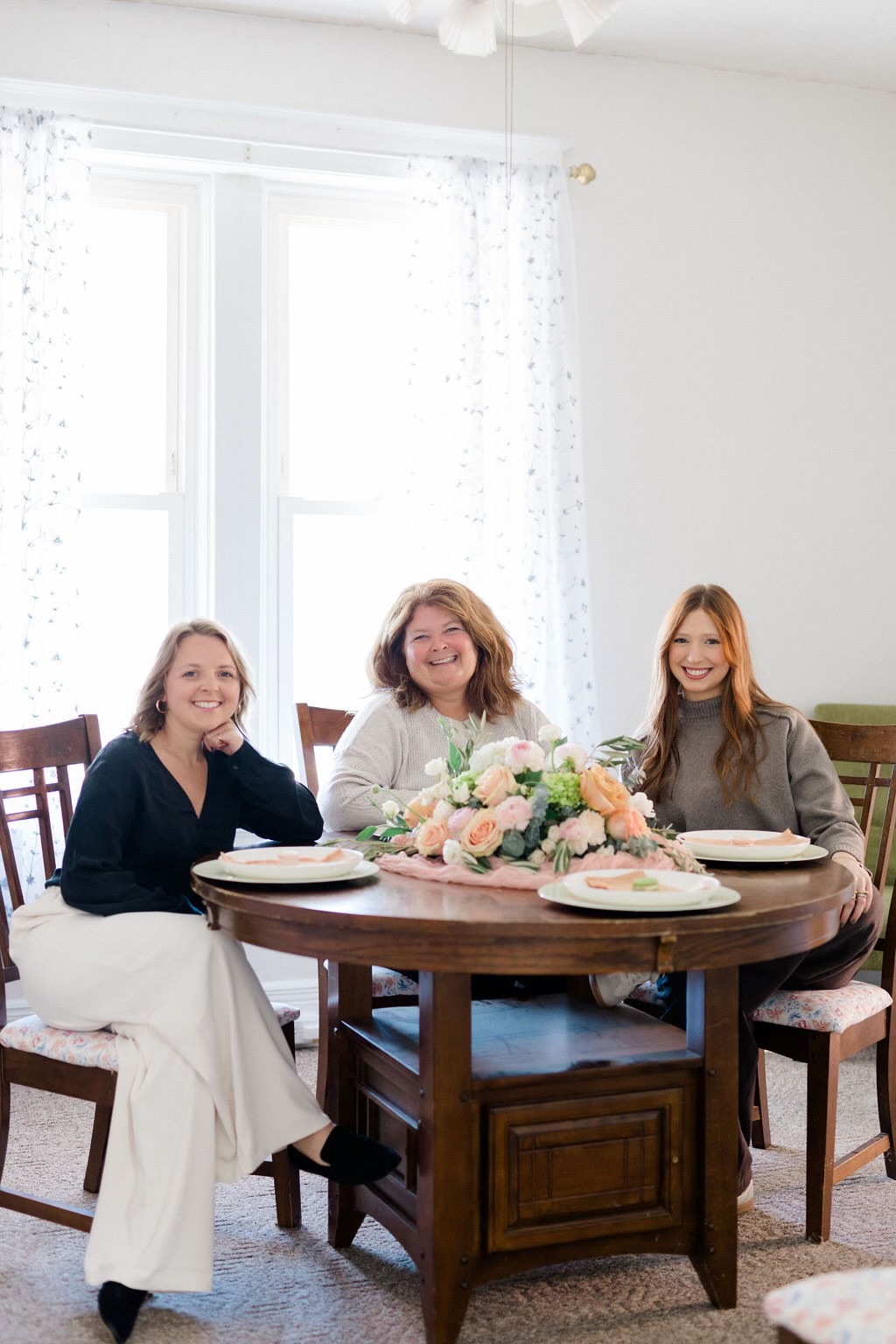 Three women sit at a round wooden table with a floral centerpiece in a bright room with large windows and white curtains.