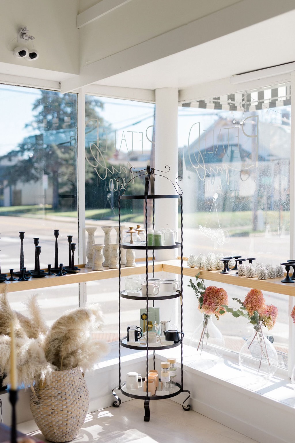 Interior view of a bright room with large windows, a black metal shelf, and decorative items, including flowers and candles.